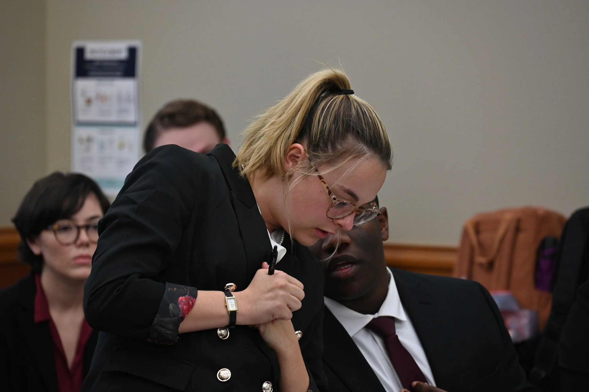 Young woman with blonde hair tied back, wearing glasses and a black jacket, engaged in a discussion or note-taking in a meeting or classroom setting with other people sitting around her.