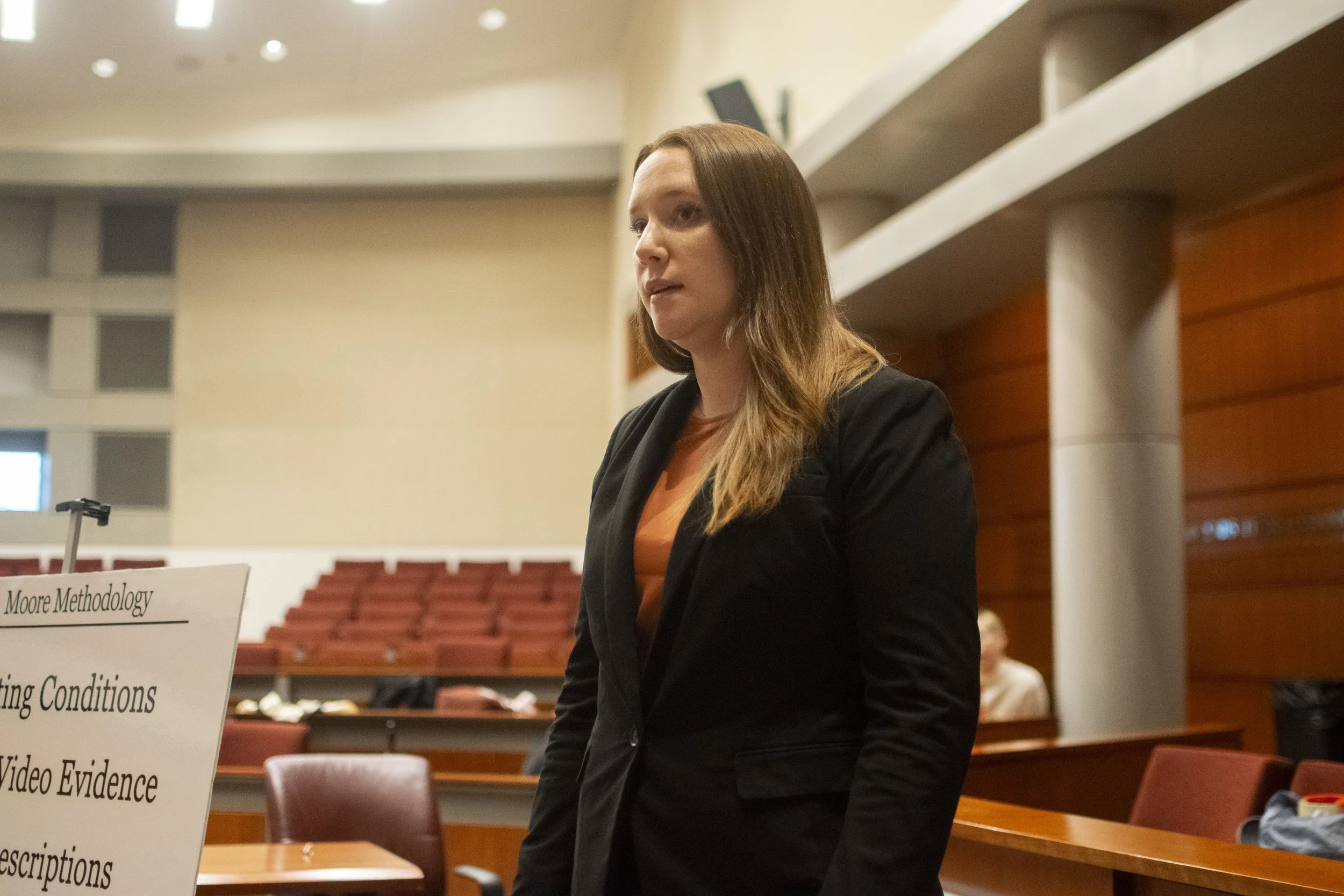 A woman with long brown hair, wearing a black blazer and orange shirt, standing in a room with wooden panels and red chairs, looking to her left.