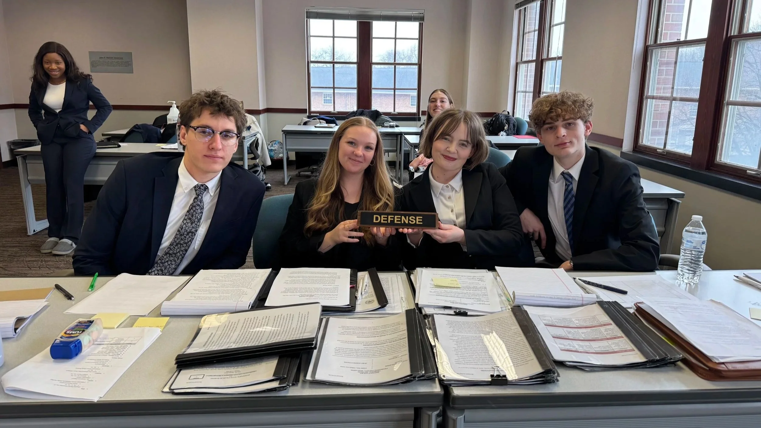 A group of six young people in professional attire in a classroom or conference room. Four are sitting at a table with documents, a water bottle, and office supplies, while two are standing behind them. The person in the middle of the seated group holds a sign that says "DEFENSE." The setting has large windows and natural lighting.