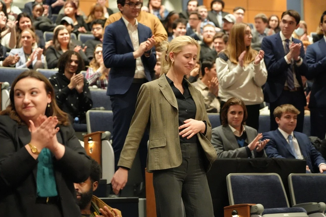 A woman with blonde hair in professional attire stands with her hand on her stomach in front of a seated audience in an auditorium, with people applauding.