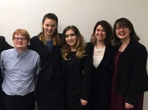 Five young women and one young man standing together in front of a plain white wall, smiling.