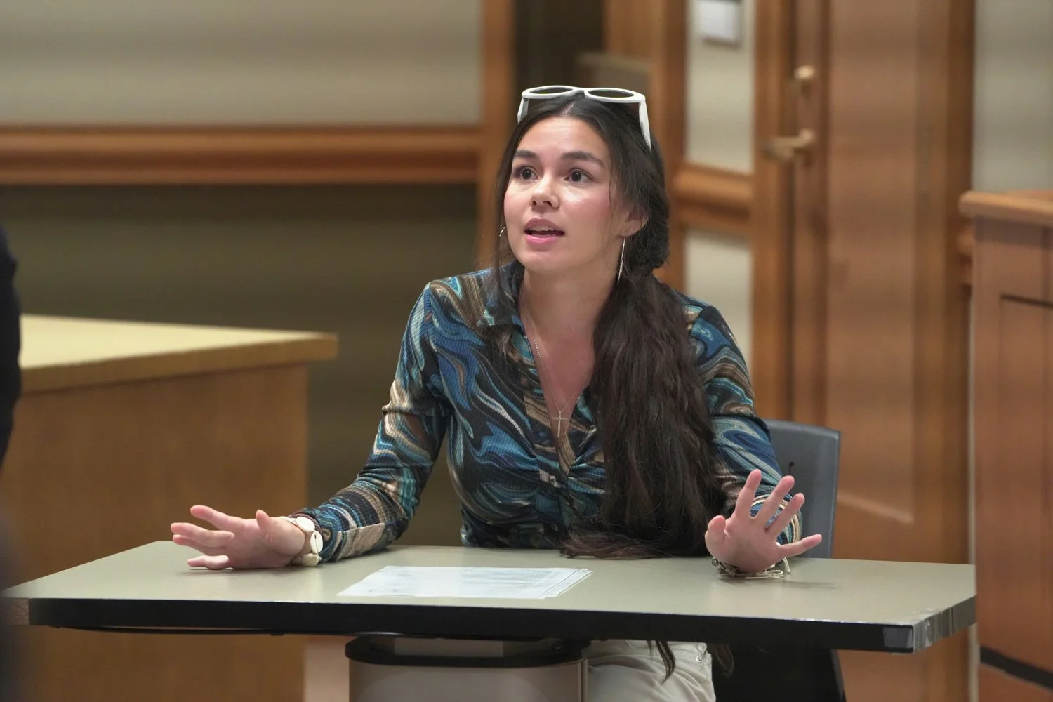 A woman with long dark hair, wearing sunglasses on her head and a colorful patterned blouse, is sitting at a table in a courtroom, gesturing with her hands while speaking.