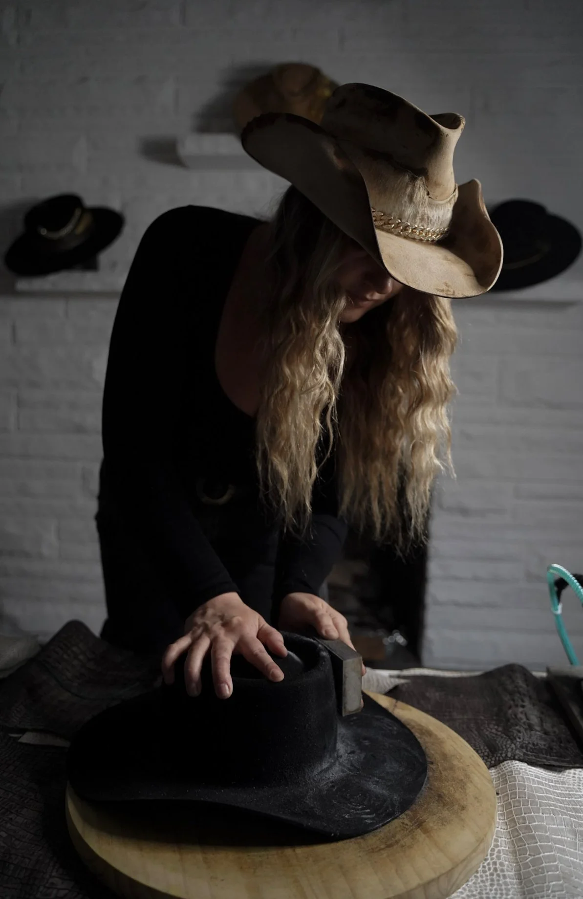 Woman wearing a cowboy hat with a chain, pressing a black hat with a hammer on a wooden surface. Hats are hanging on a white brick wall in the background.