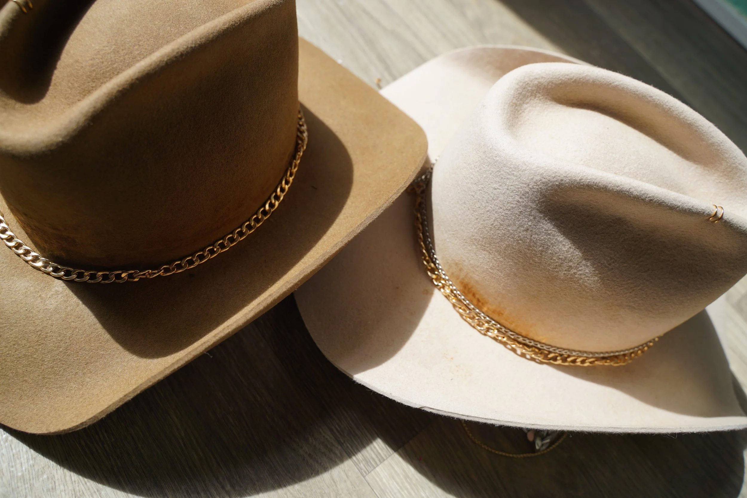 Two wide-brimmed hats, one brown and one cream, both with gold chain embellishments, are shown in the image. The hats are placed on a wooden surface with sunlight casting shadows.