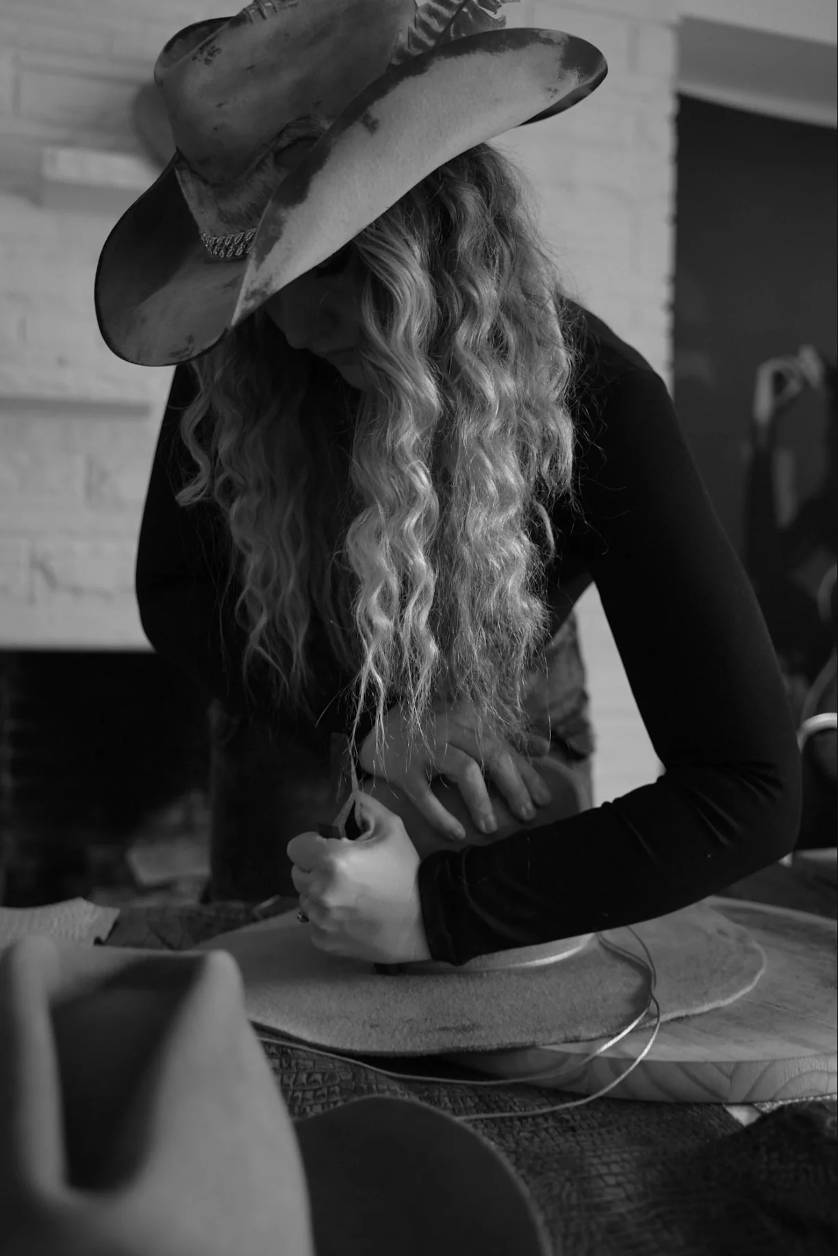 A woman with long curly hair wearing a wide-brimmed hat and black long-sleeve shirt, working on a craft project at a table.