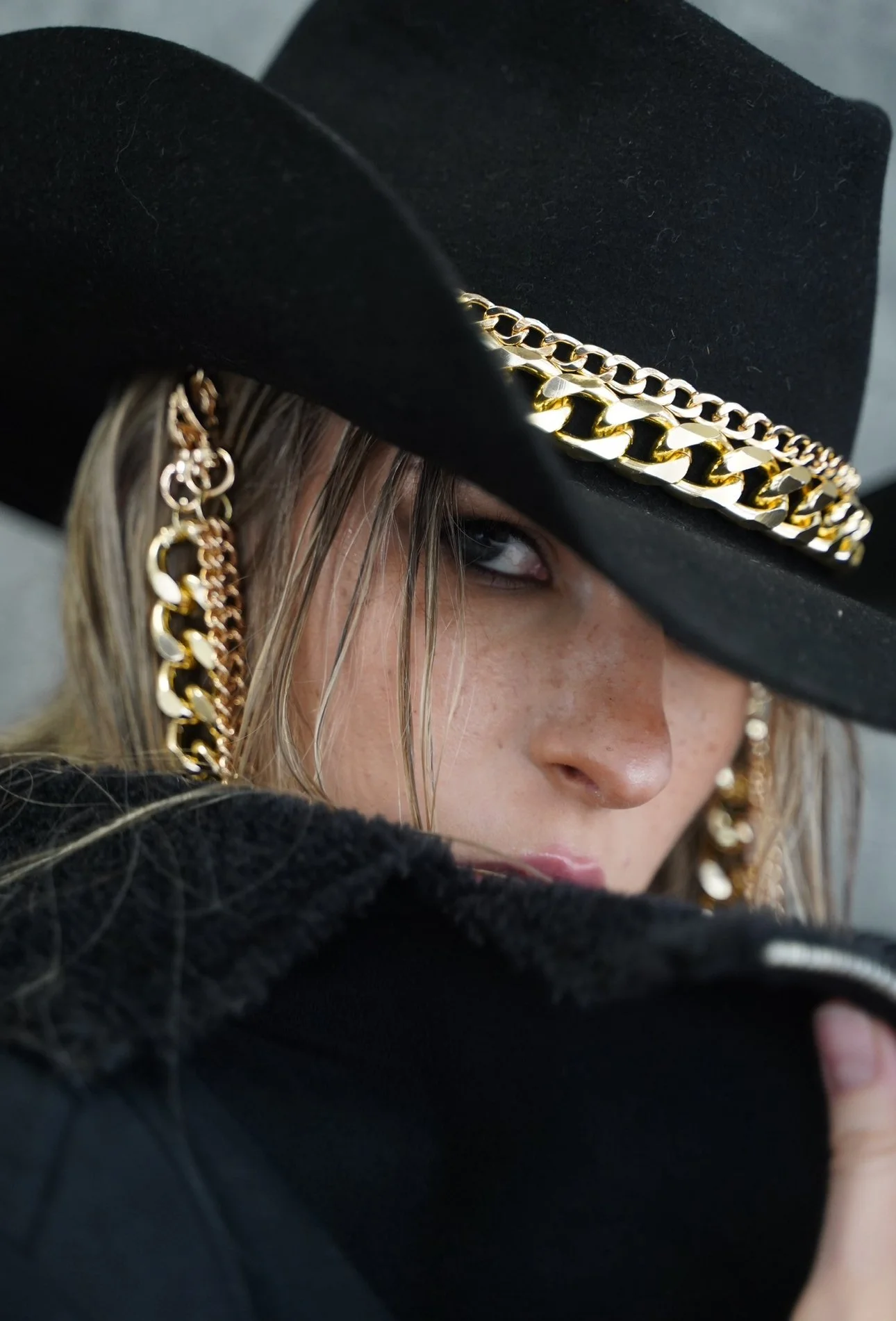 A woman wearing a black hat with gold chains, large gold chain earrings, and a black jacket, partially covering her face with her collar.