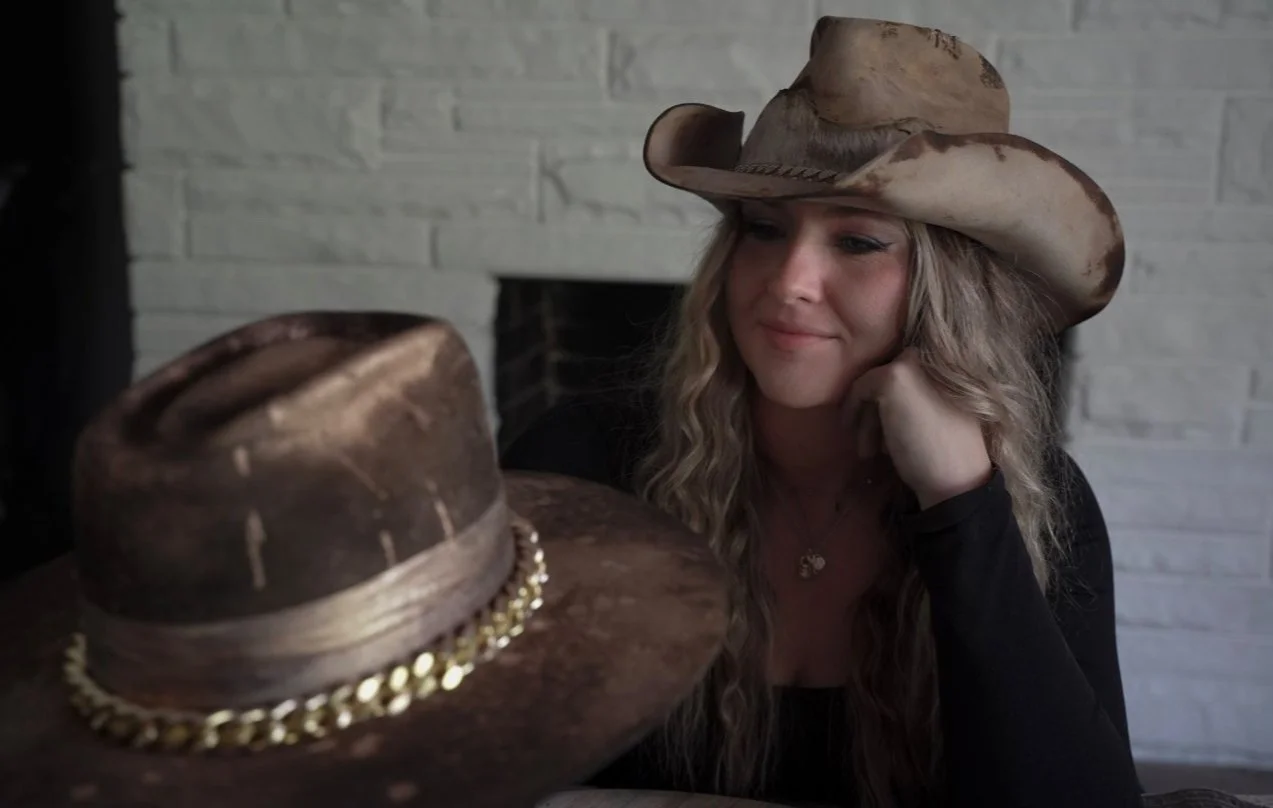 A woman with long wavy hair wearing a cowboy hat, looking at a distressed hat with a chain decoration, in front of her. She is sitting indoors against a white brick wall.
