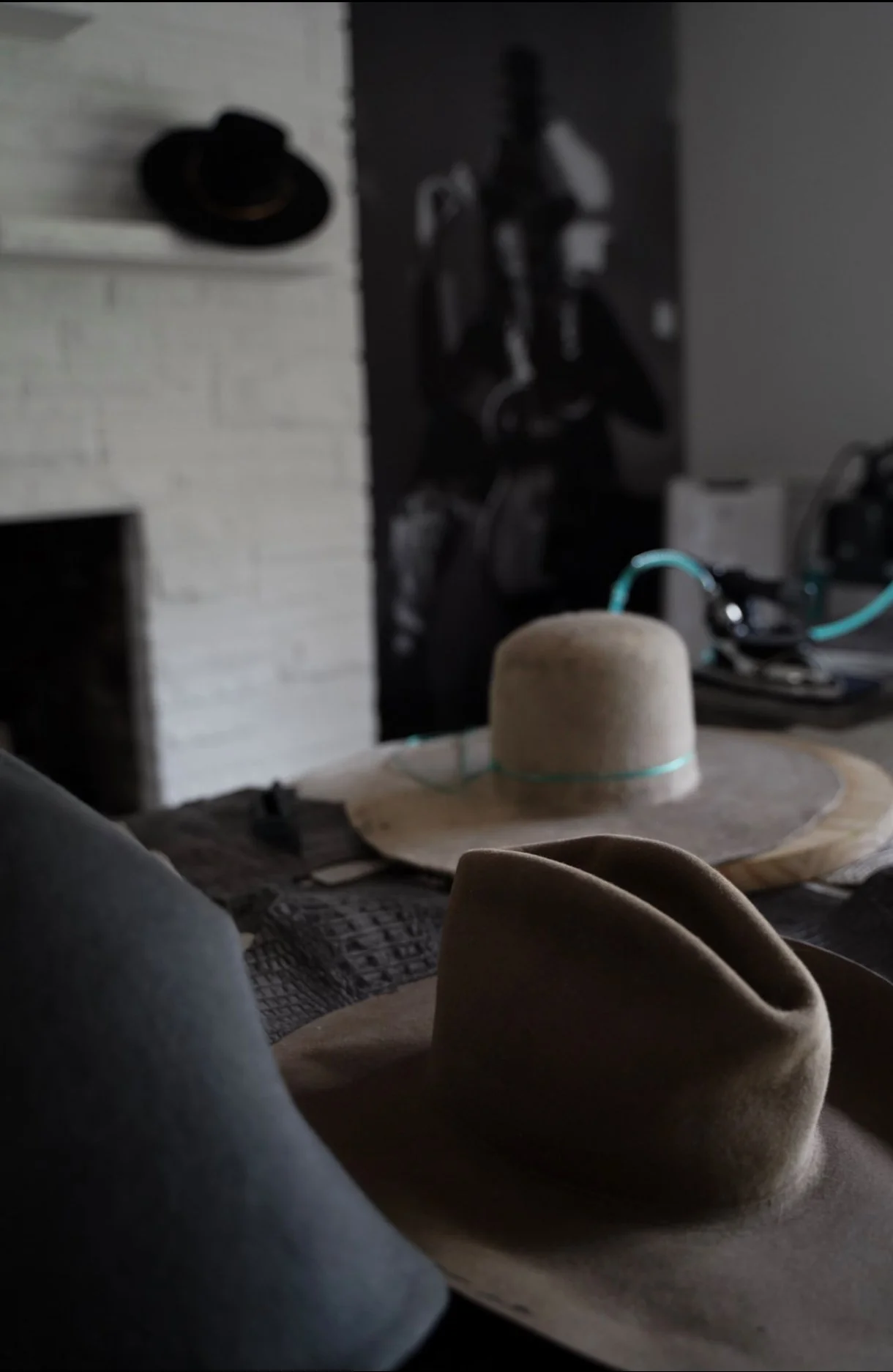 Close-up of two beige felts hats resting on a wooden surface with a black hat hanging on a white brick wall in the background.