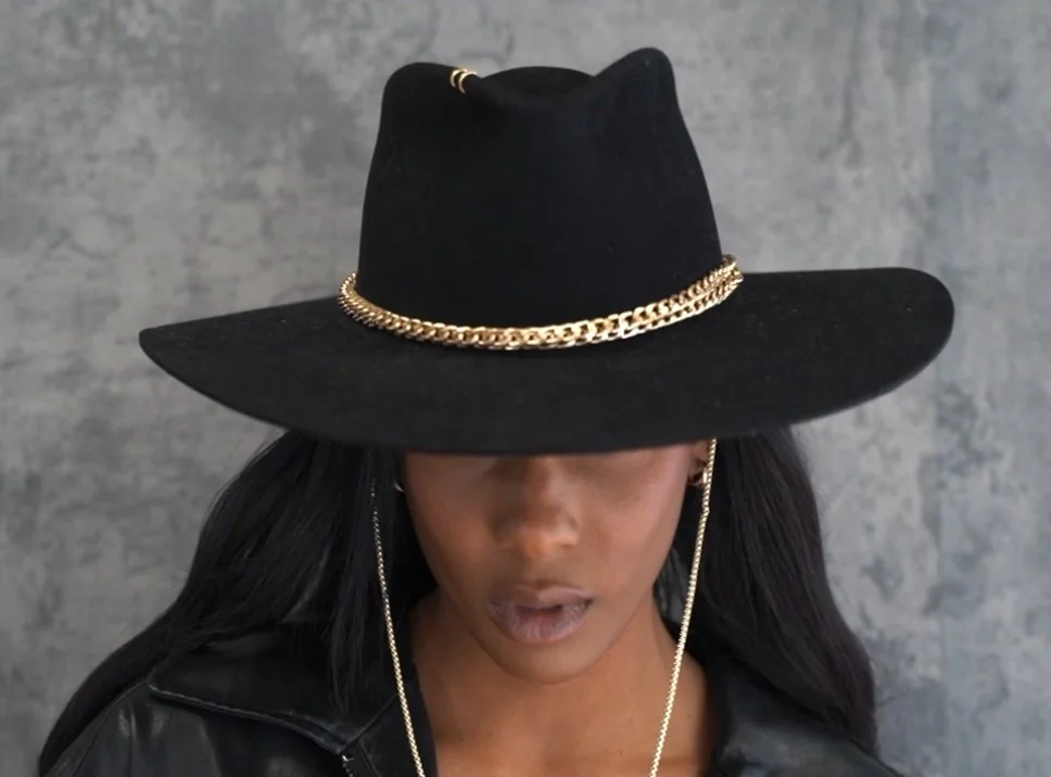 A woman wearing a black wide-brimmed hat with gold chains, a black leather jacket, and jewelry.