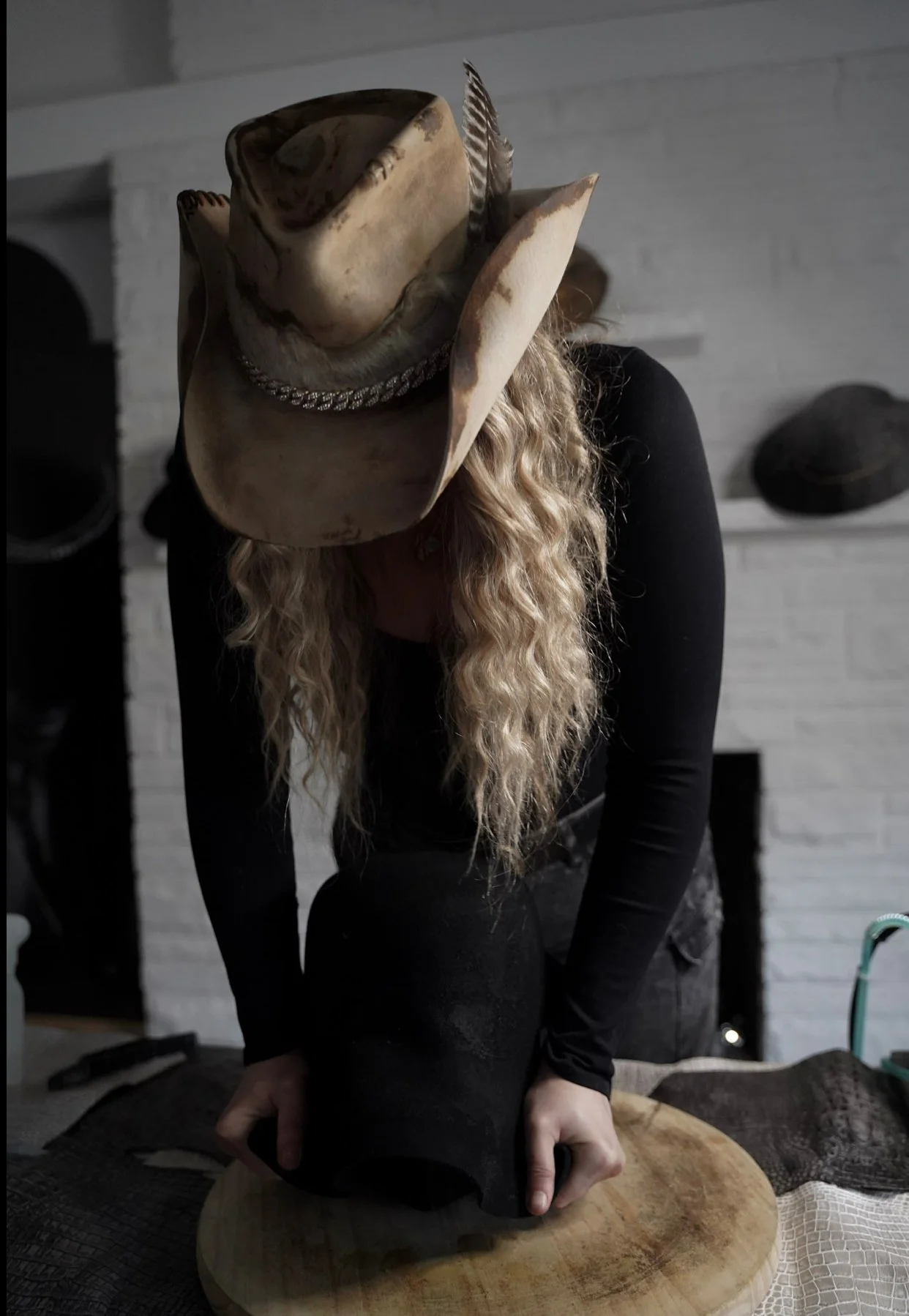 Person wearing a cowboy hat with feathers and a silver chain, bent over a black object on a wooden surface, in front of a white brick wall with hats on shelves.