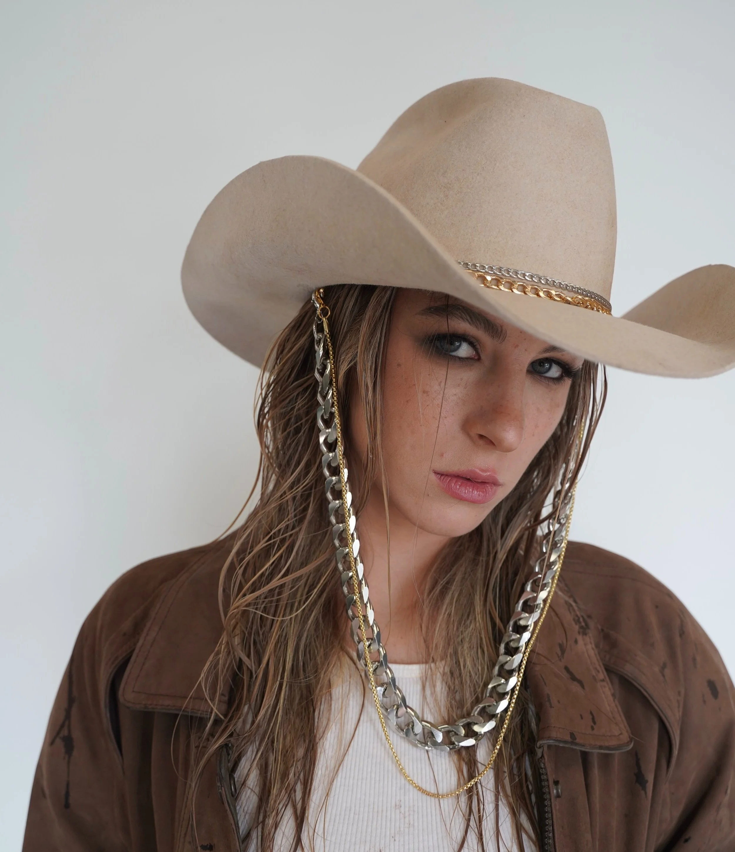 A woman wearing a beige cowboy hat with silver and gold chains draped around it. She has wet, wavy hair, blue eyes, and is dressed in a white ribbed shirt and brown jacket, standing against a plain light background.