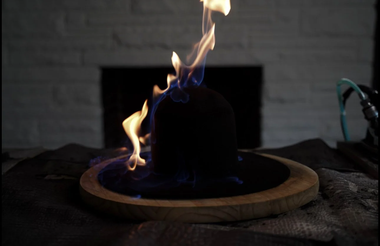 A small black hat on a wooden plate, with flames and blue smoke burning on top, in front of a dark brick wall.