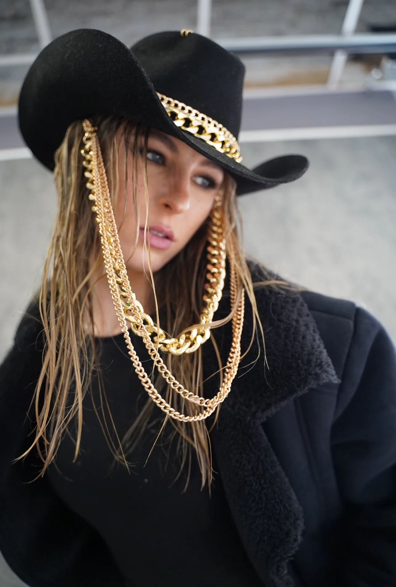 A woman wearing a black cowboy hat decorated with a gold chain and layered gold necklaces, with wet hair and a black coat.