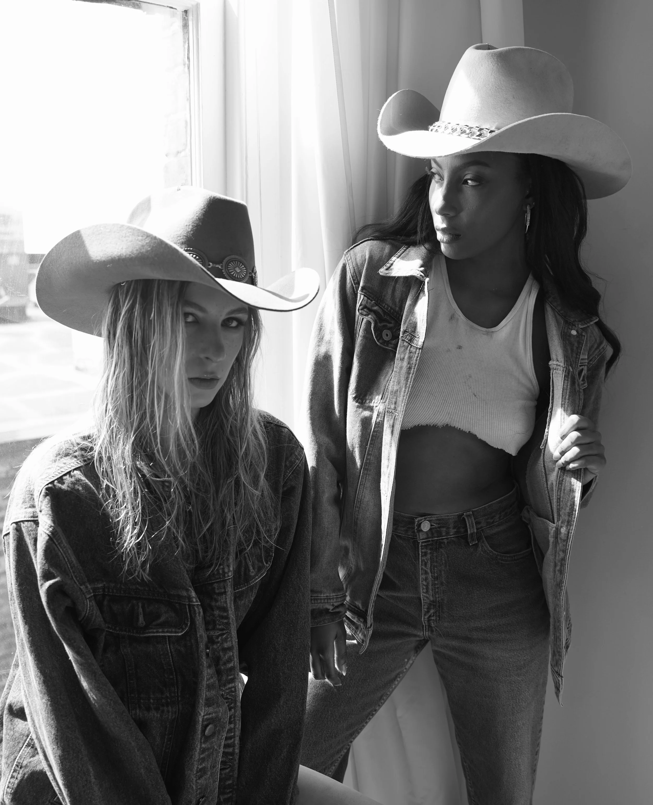 Two women wearing cowboy hats and denim jackets, standing by a window in black and white