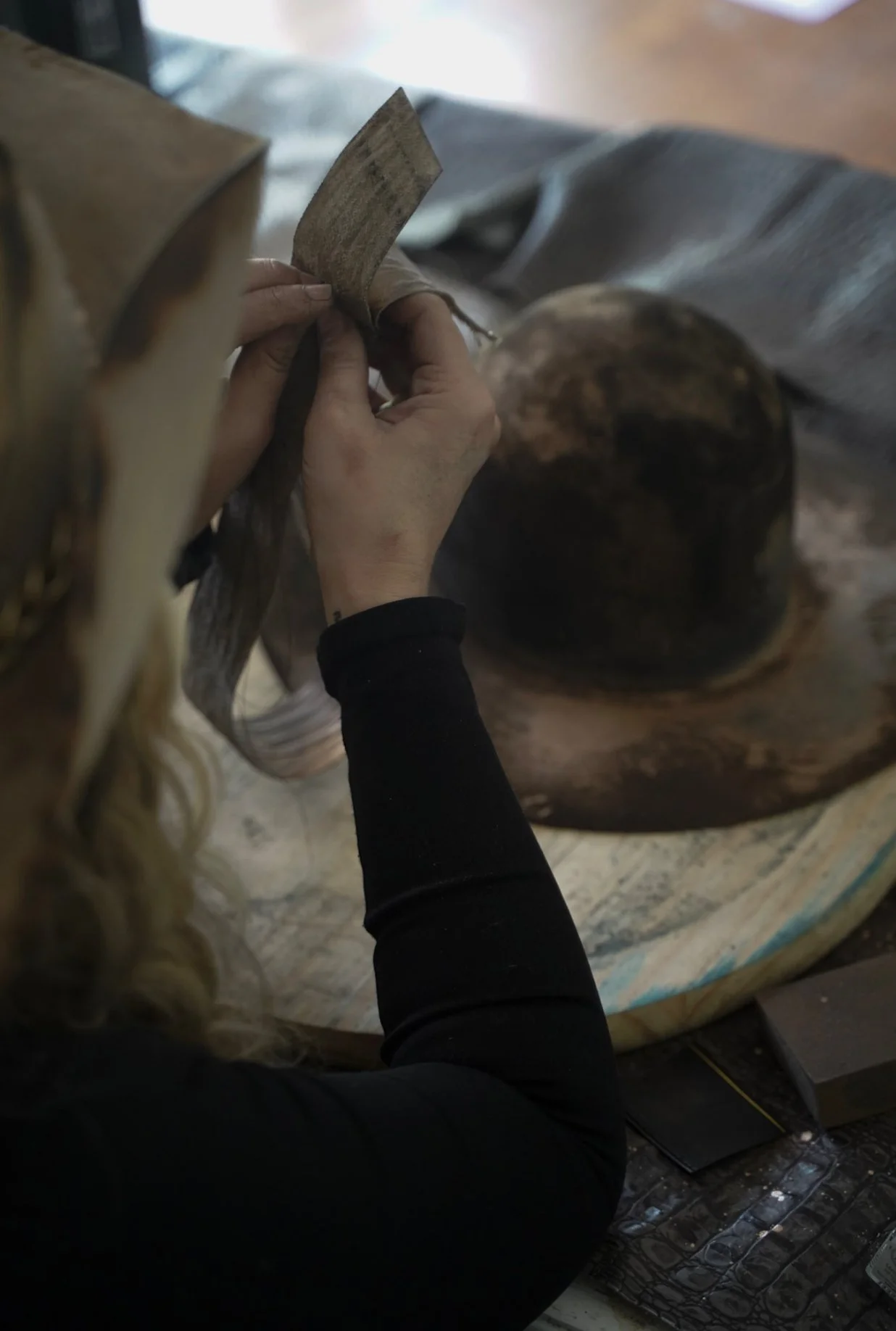 A person with curly blonde hair working on a piece of leather at a wooden workbench, with a worn hat nearby.