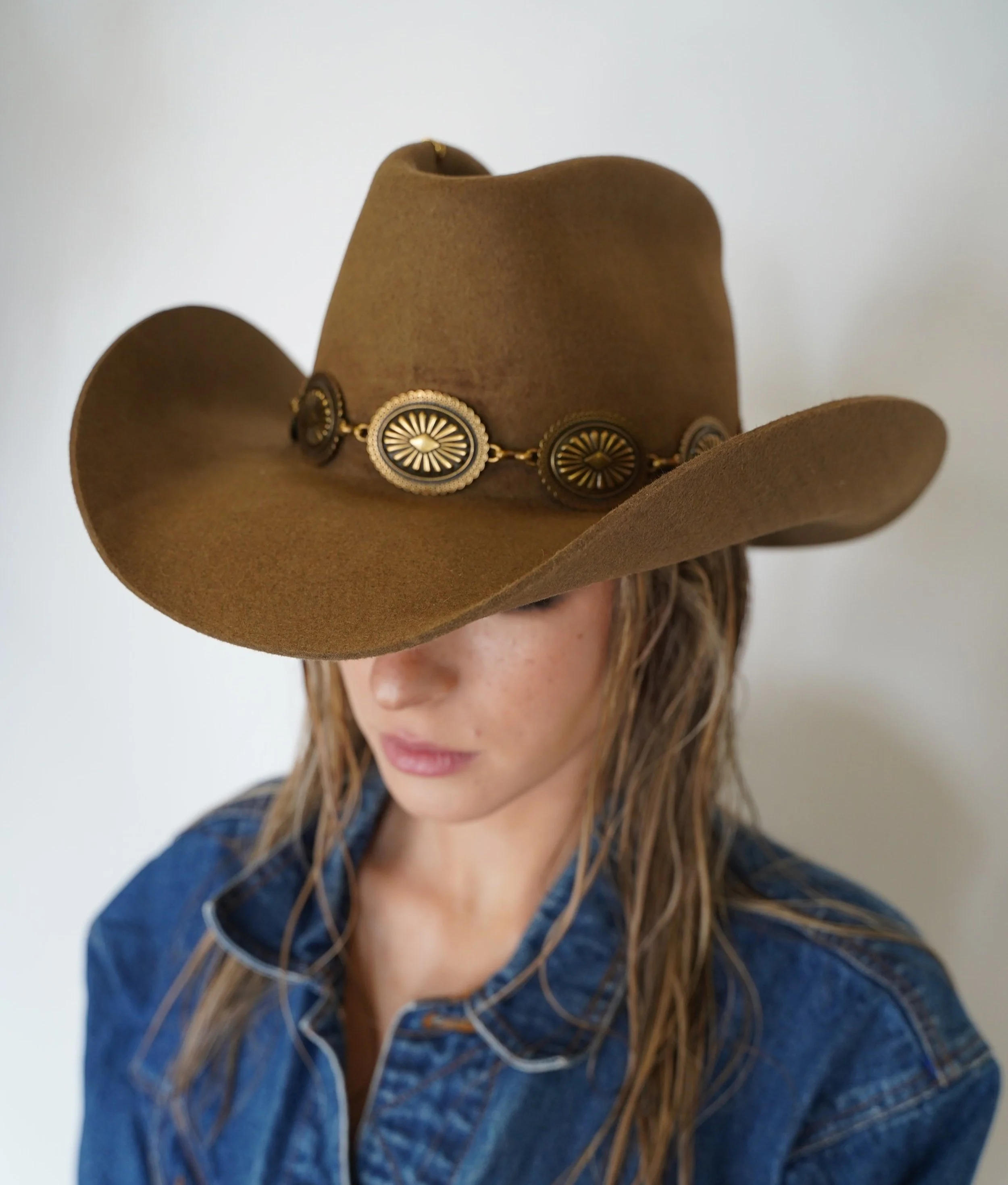 A woman wearing a brown cowboy hat with a decorative band and a blue denim shirt.