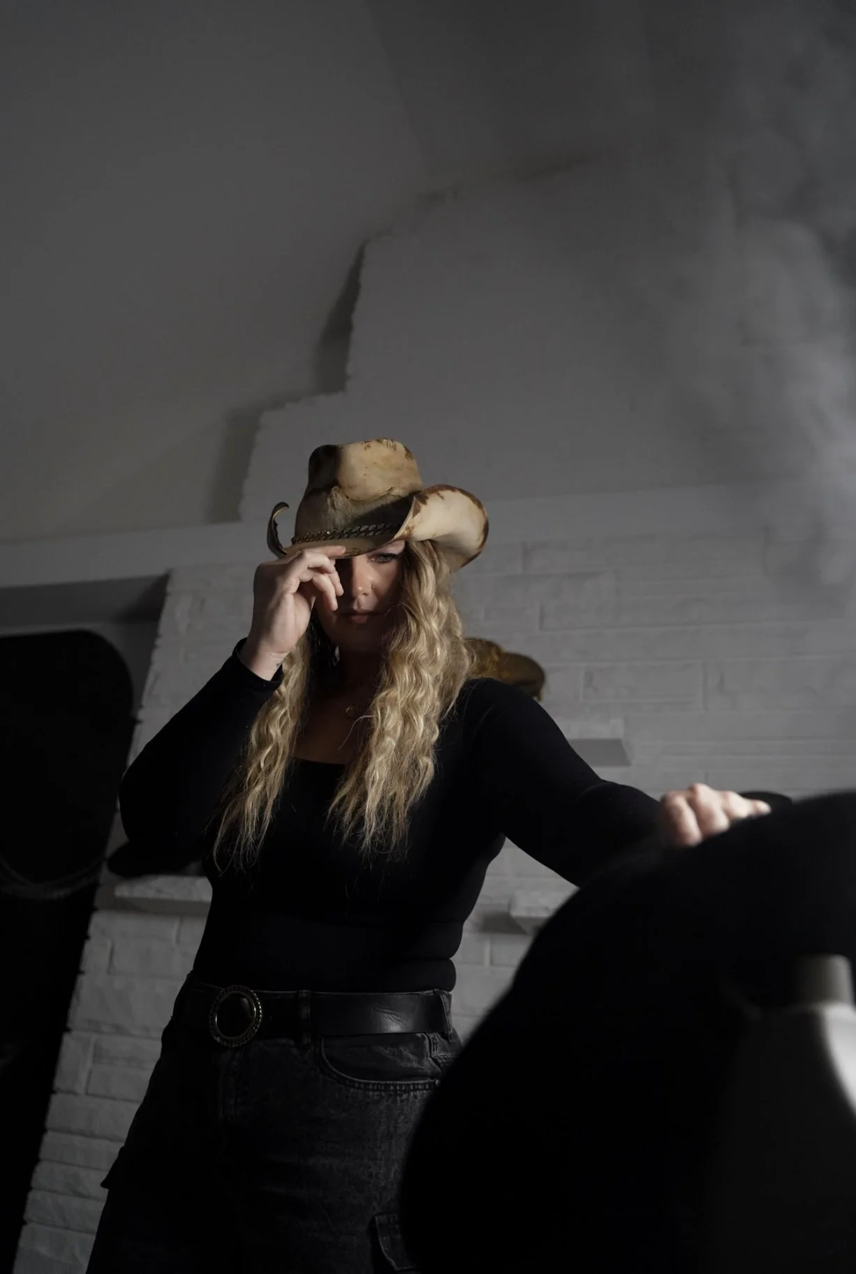 A woman wearing a cowboy hat and black outfit, looking down as she touches a large object, with a white brick wall and ceiling in the background.