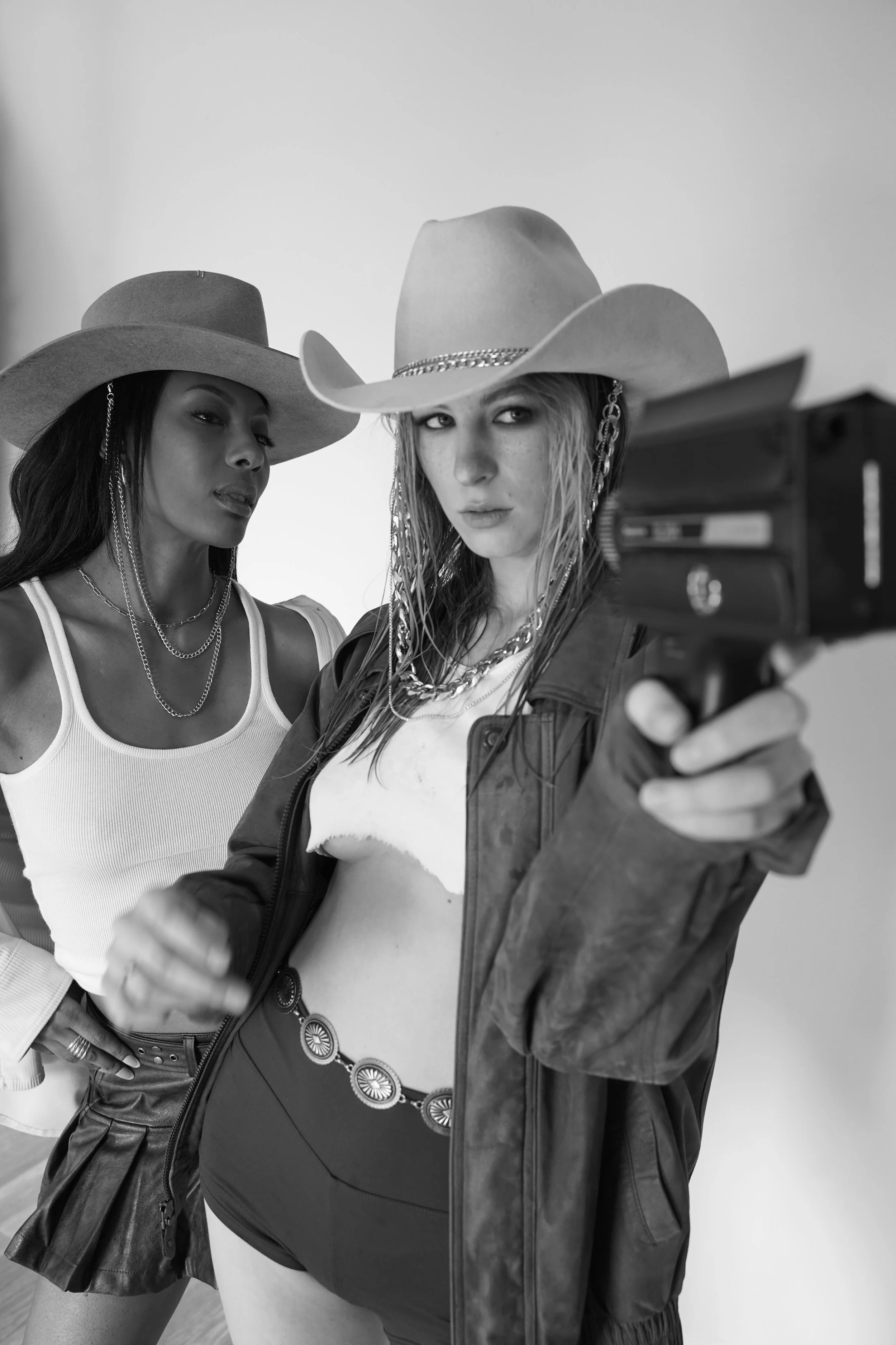 Two women in cowboy hats and chain necklaces taking a selfie with a gun, one in a leather jacket, in a black-and-white photo.