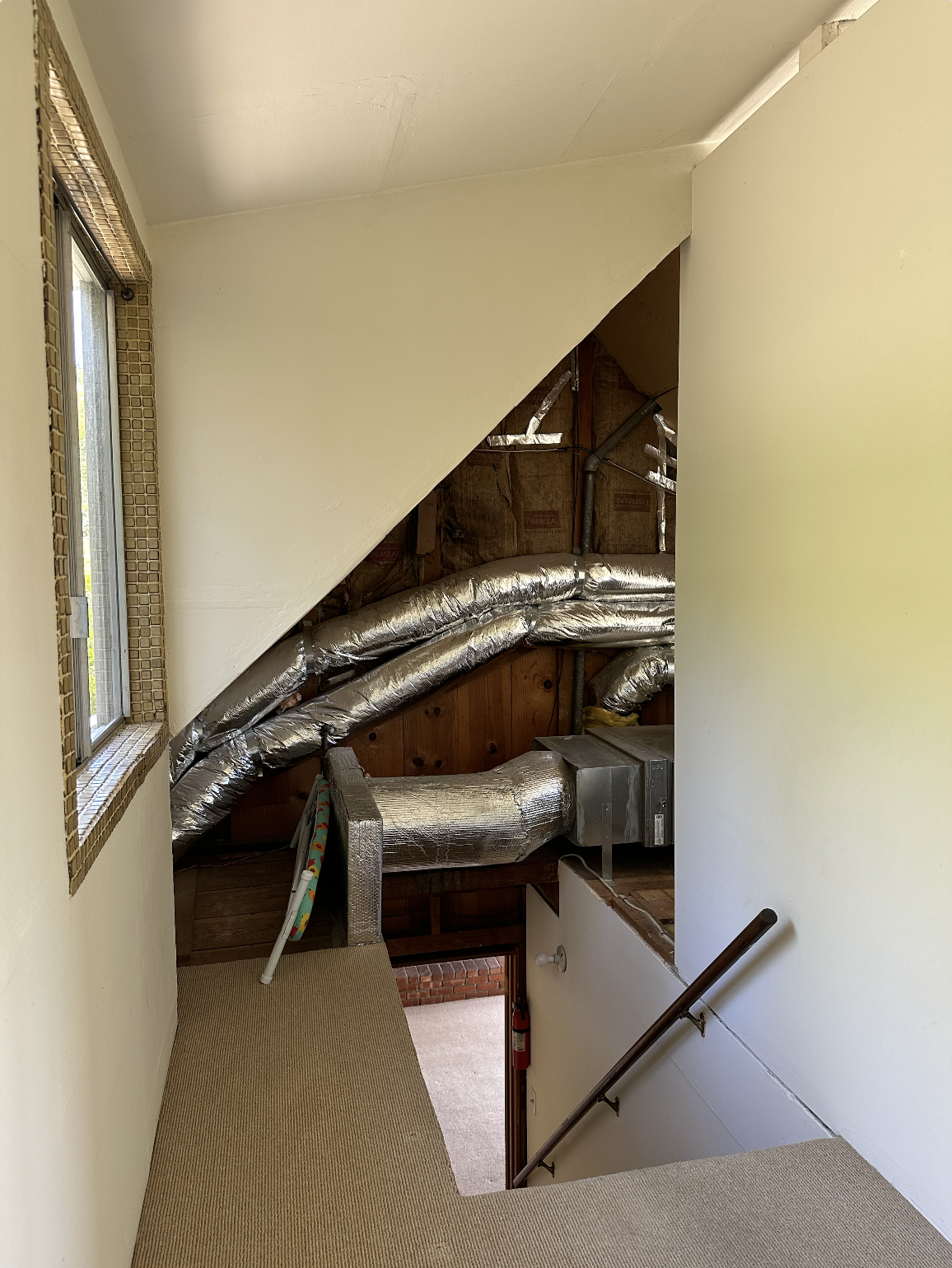 Interior view of an attic or space under a staircase, showing exposed ductwork & insulation, with a window on the left and a staircase with a handrail on the right.