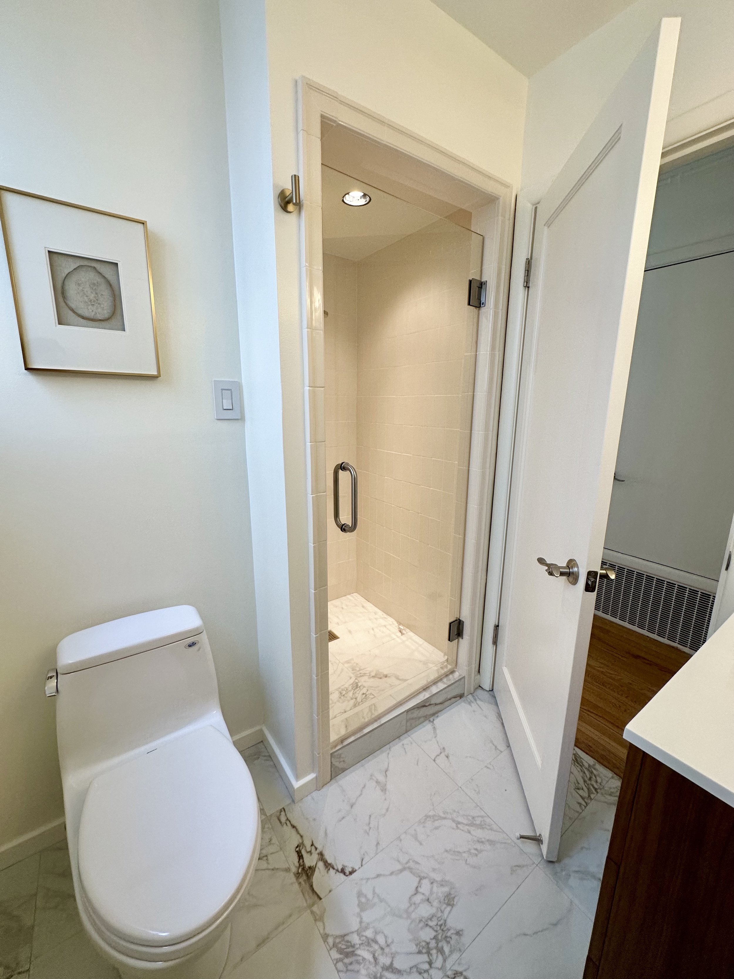 A small bathroom with a white toilet, a framed artwork on the wall, and a glass shower with beige tiles and a marble floor. The door is open to a room with wood flooring.