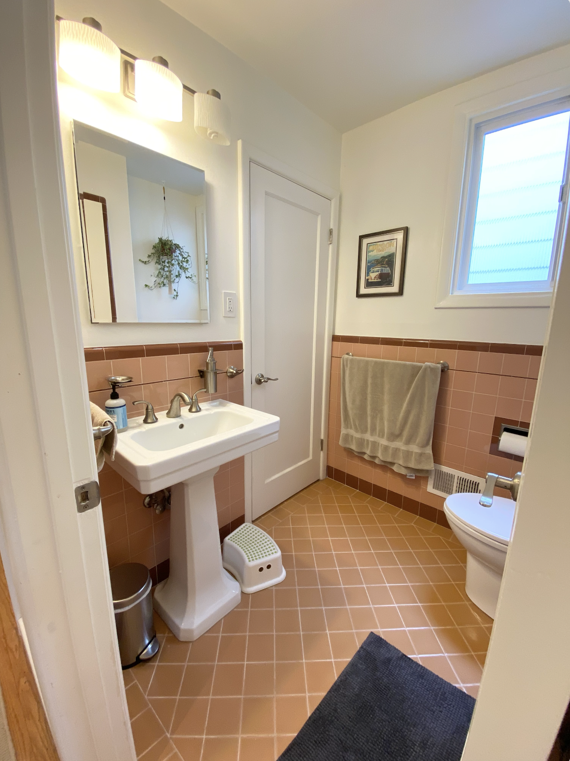 A small bathroom with peach-colored tiled walls and floor. It features a pedestal sink with a mirror above, a towel hanging on the wall, a small window, and a toilet. There are various small items and a trash can next to the sink.