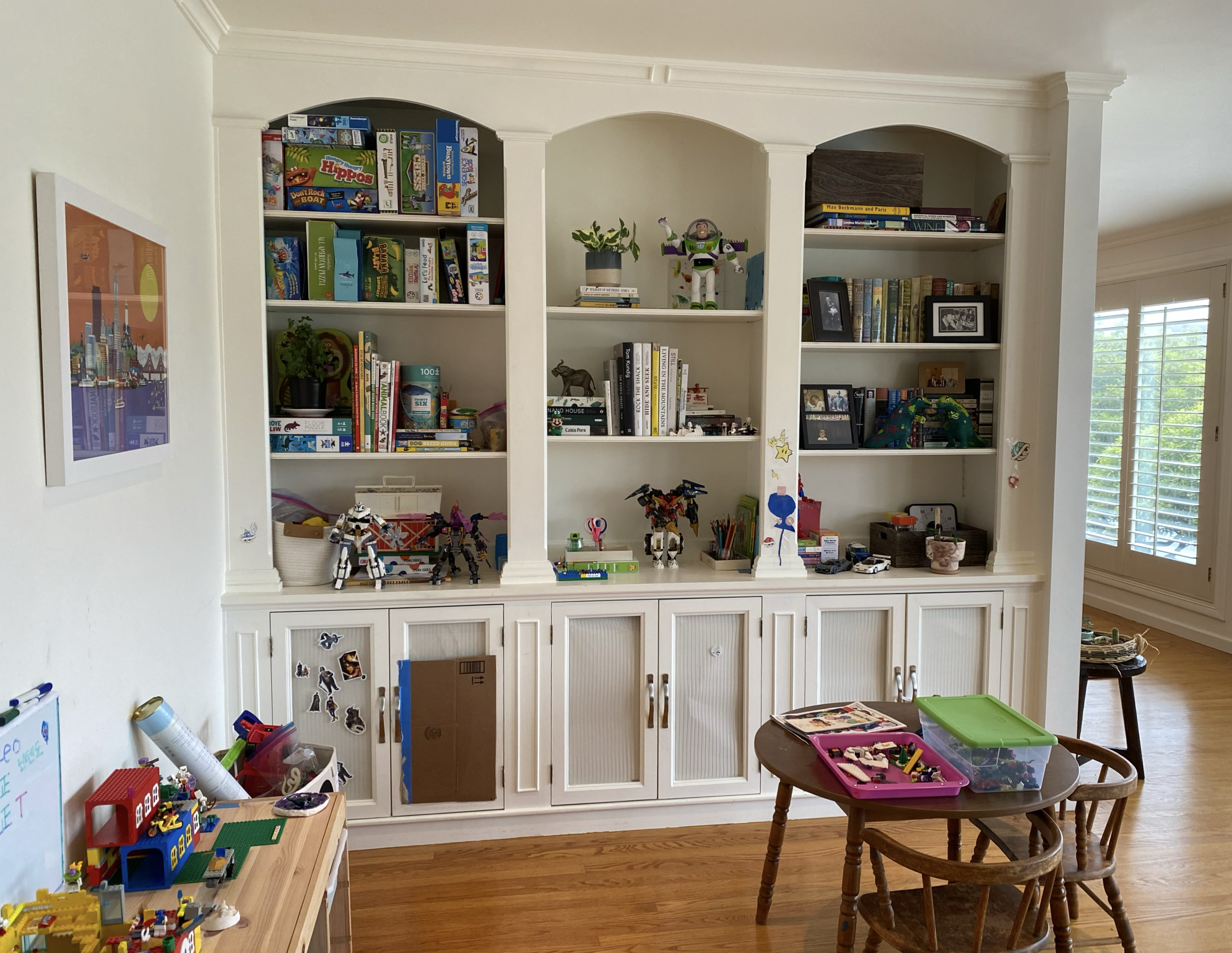 A living room with a large built-in white bookshelf filled with books, toys, and framed photos. Lego sets and toys are on the lower shelf and on a table in the forefront. There is a small round wooden table with Lego pieces, a green container, and a toy dinosaur. A wall with Korean letters and drawings is partially visible on the left, and a window with white shutters is on the right.