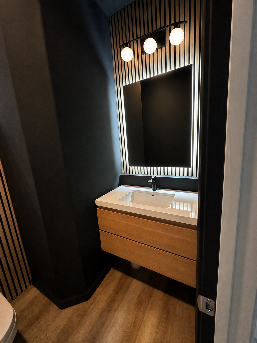 Modern bathroom vanity with a black faucet, wooden cabinet, white sink, illuminated mirror, and three spherical ceiling lights.