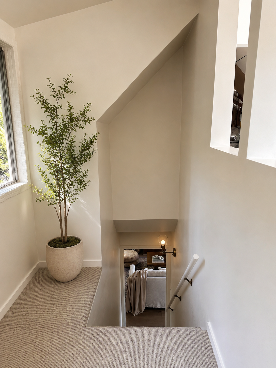 Interior view of a staircase landing with a large potted plant near a window, beige carpet, white walls, and a view of a living room with a sofa and lamp below.