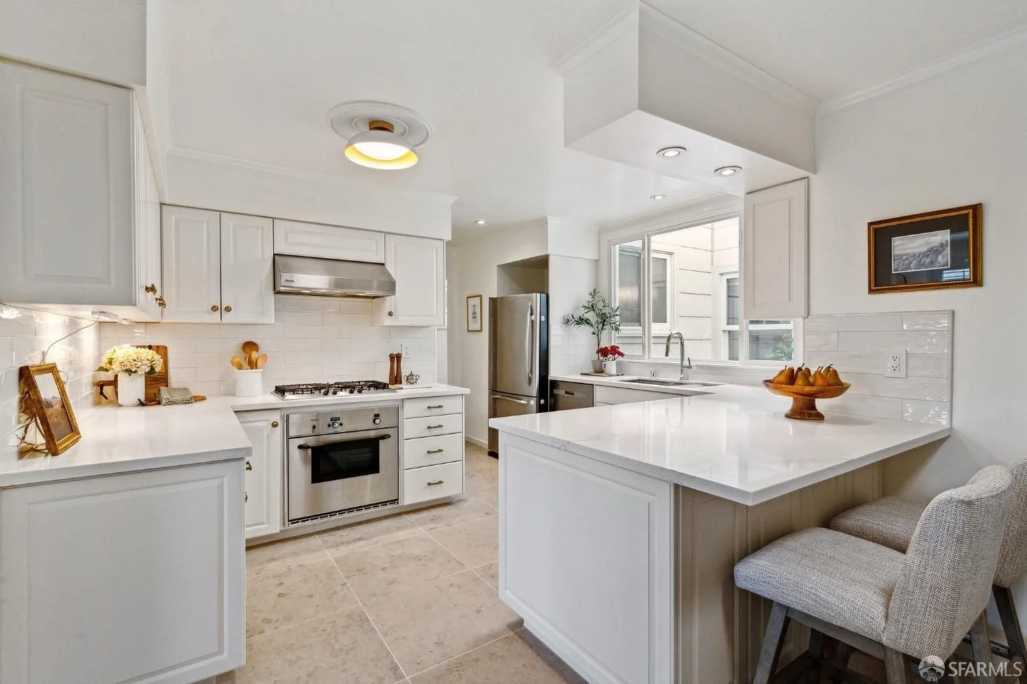 Bright white kitchen with cabinetry, stainless steel appliances, tiled floor, and a corner window. Decor includes framed photos, a flower pot, and a fruit bowl.