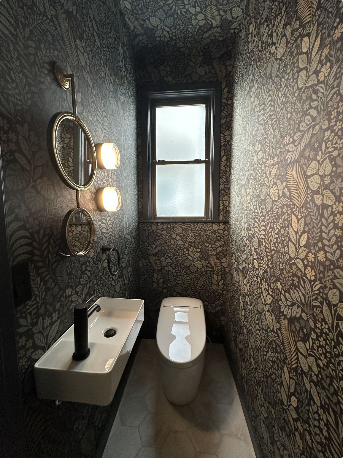 A small bathroom with dark botanical-patterned wallpaper, a frosted window, a modern white toilet, a small rectangular white sink with a black faucet, round mirrors, and two wall-mounted light fixtures.