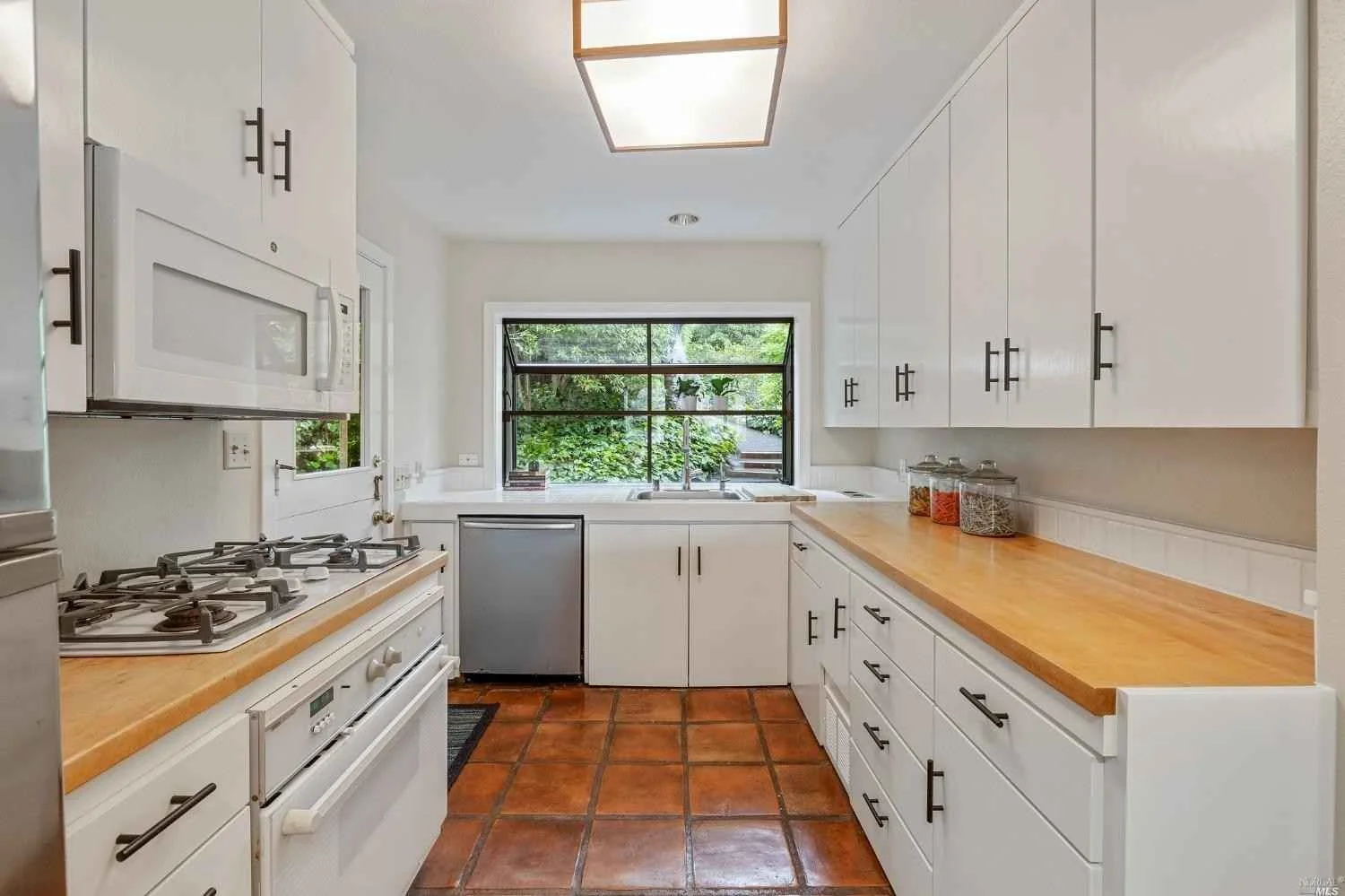 Kitchen with white cabinets, wooden countertops, terracotta tiled floor, window overlooking greenery, stove, microwave, small refrigerator, and glass jars with spices on counter.