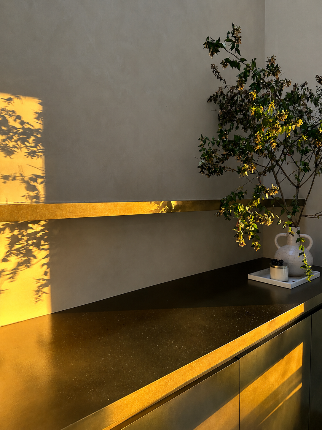 A modern interior with a dark countertop, a white vase with leafy green branches, and a tray with small items, illuminated by warm sunlight casting shadows on the wall.