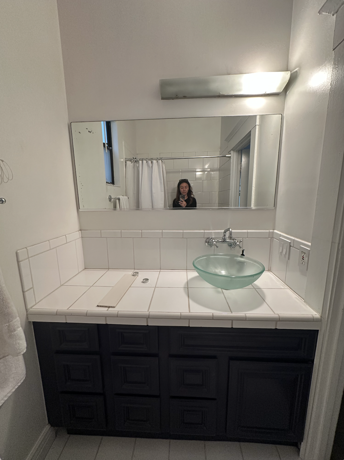 Bathroom vanity area with a large horizontal mirror, a glass vessel sink, a faucet above the sink, a dark cabinet below, and a white tiled countertop. In the mirror, a woman taking a selfie, a window, a shower curtain, and a towel are visible.