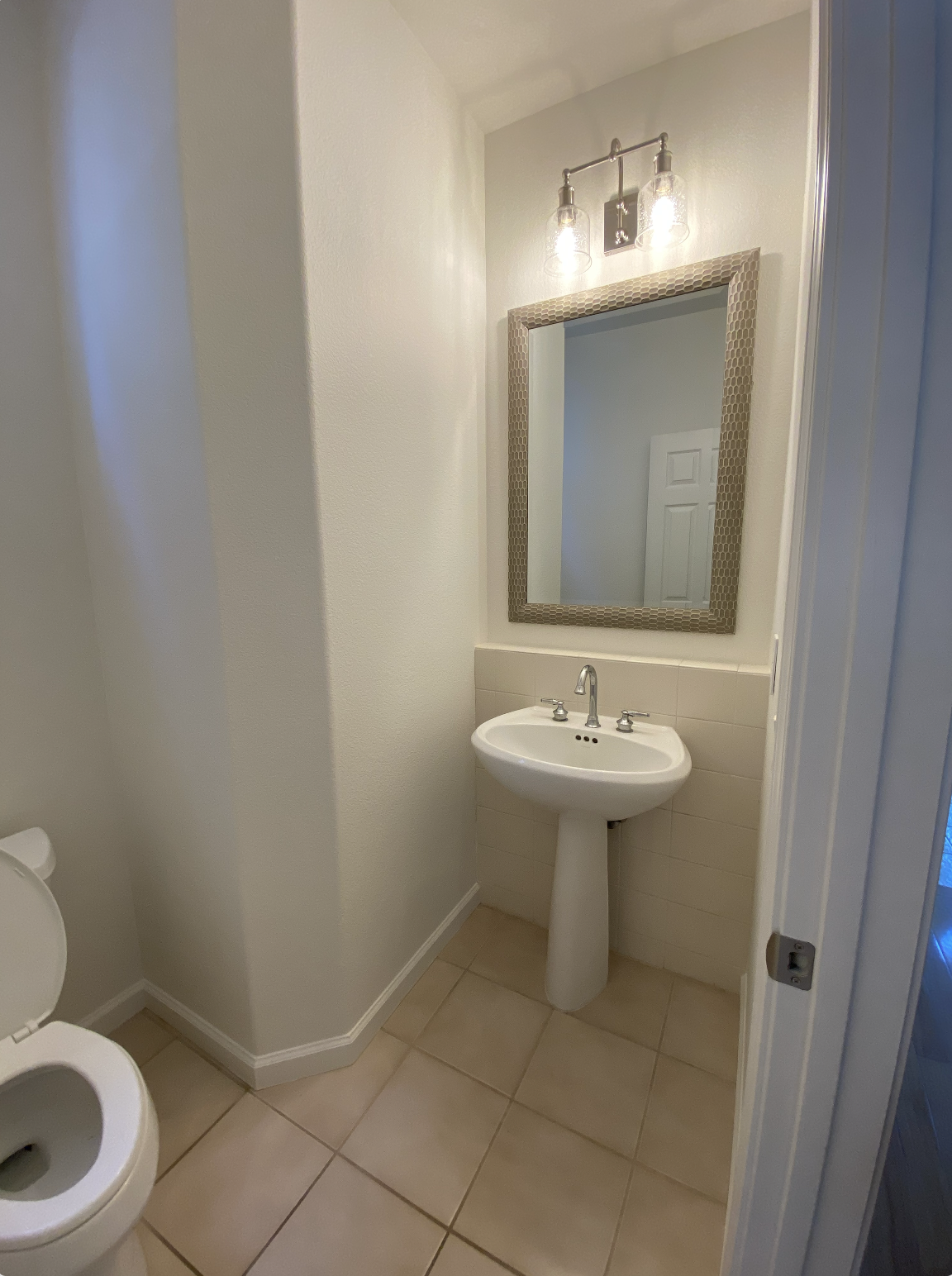 Small bathroom with a pedestal sink, a mirror with a decorative frame, and a light fixture above the mirror. Part of a toilet is visible on the left, and the room has tiled floor and walls painted in a light color.