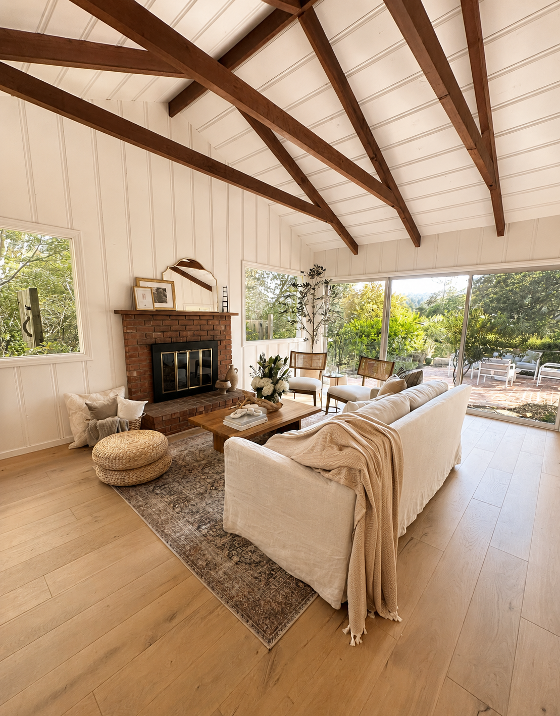 Bright living room with white walls, wooden ceiling beams, and large windows and sliding glass door showing outdoor greenery. Contains a cream-colored sofa with a beige throw, a wooden coffee table with a floral centerpiece, and various chairs and decorative items, including a brick fireplace.