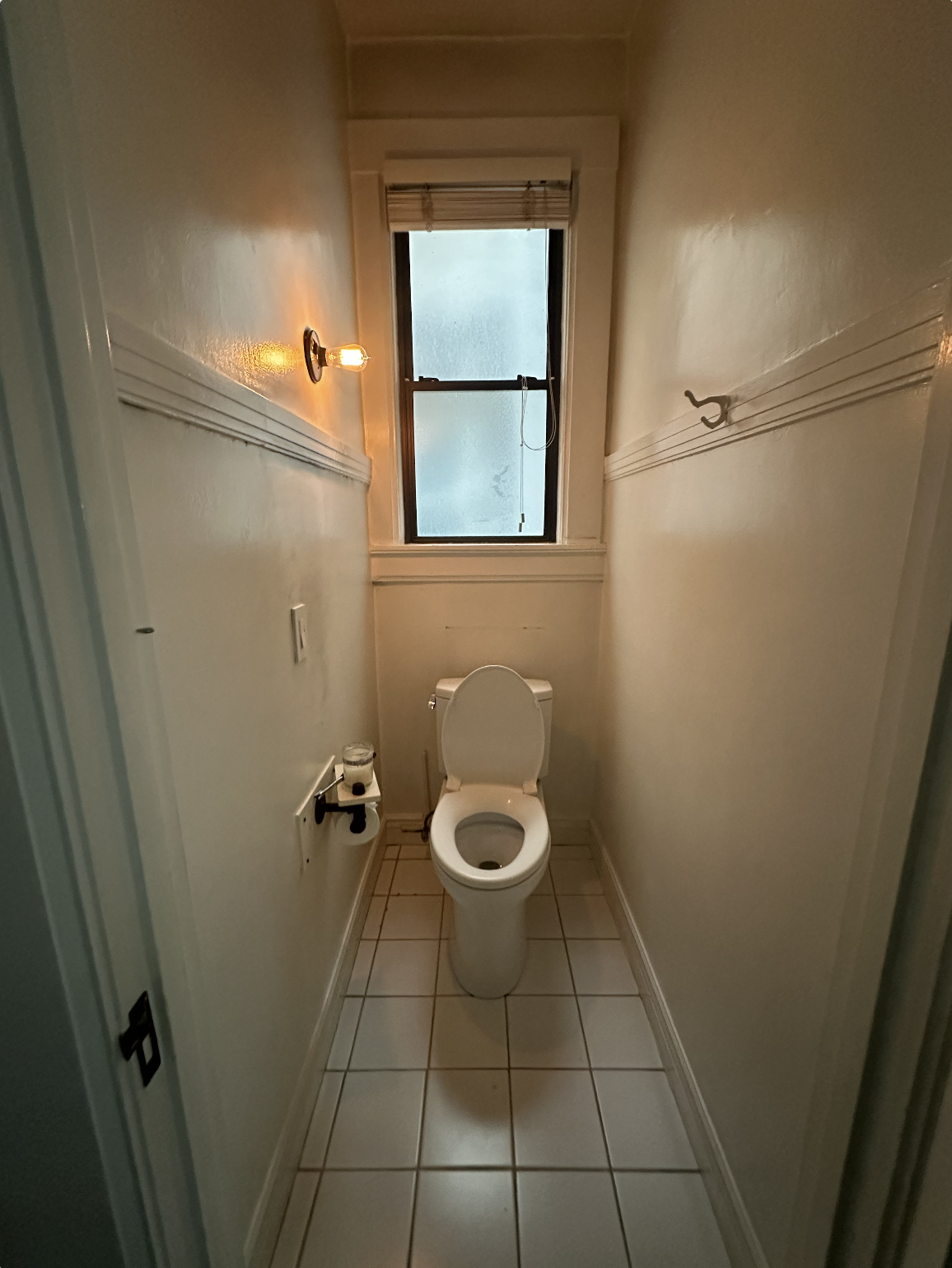 A small, narrow bathroom with beige tile flooring, cream-colored walls, a toilet, a window at the end, and a small wall-mounted light fixture.