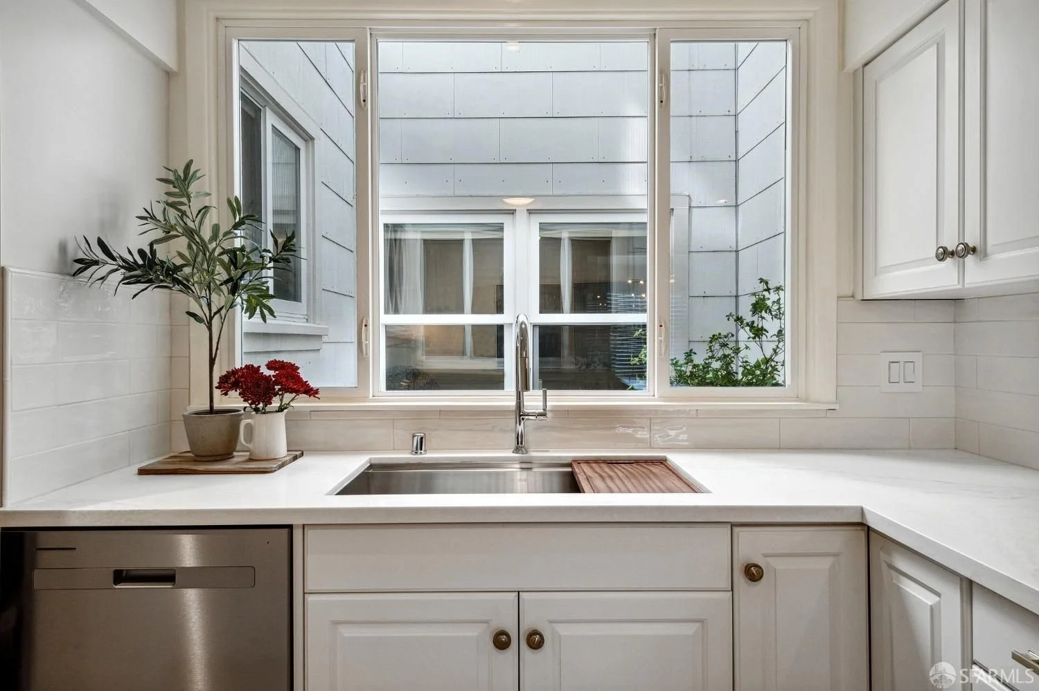 Kitchen sink area with a window, potted plants, white cabinets, marble countertops, and a dishwasher.
