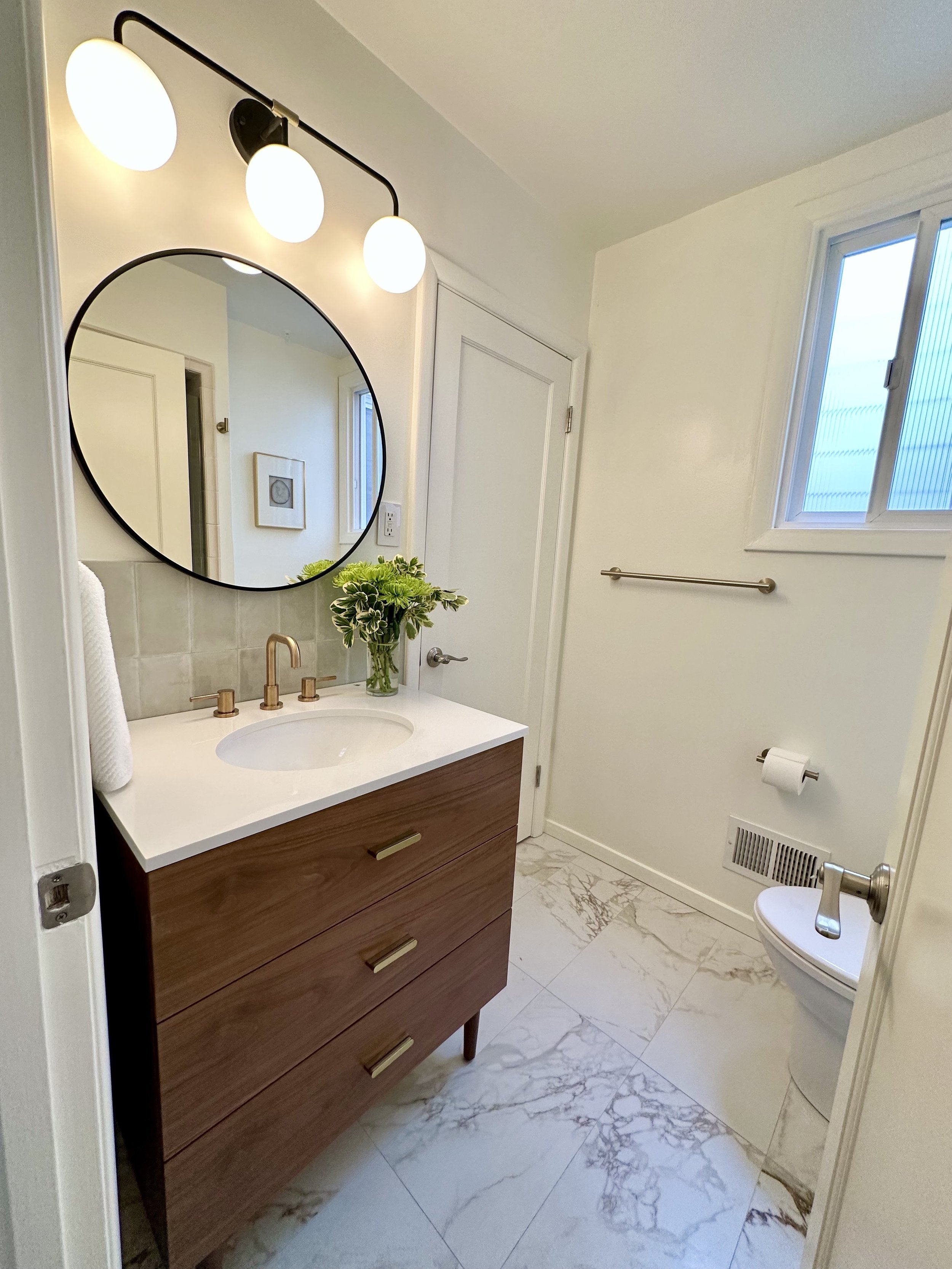 A modern bathroom with a wooden vanity, round mirror, white countertop, green plant, wall light fixtures, and a window. There is a toilet and a towel rod on the wall.