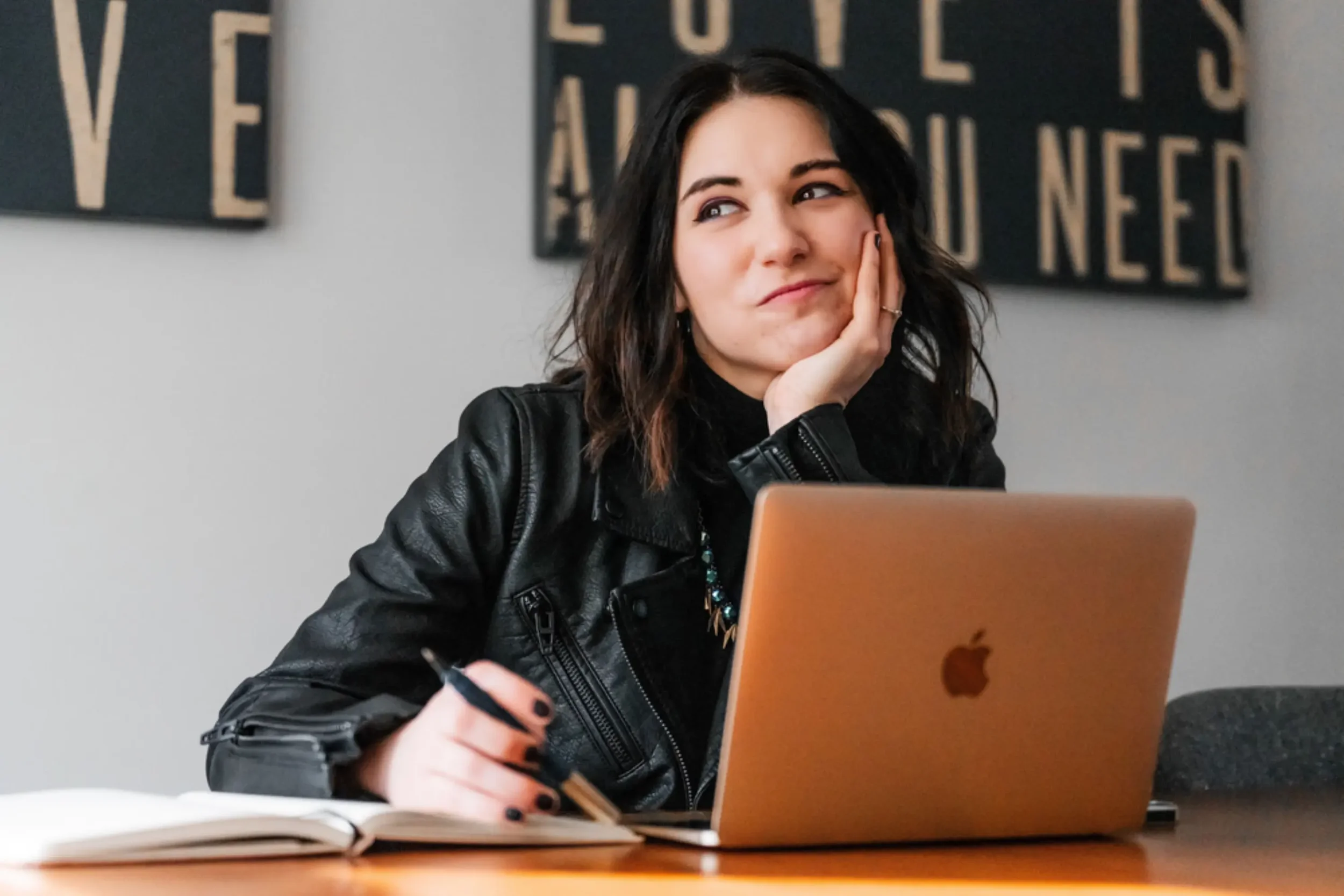 Young woman with dark wavy hair wearing black leather jacket sitting at table with laptop and notebook, resting her head on her hand in a thoughtful pose.