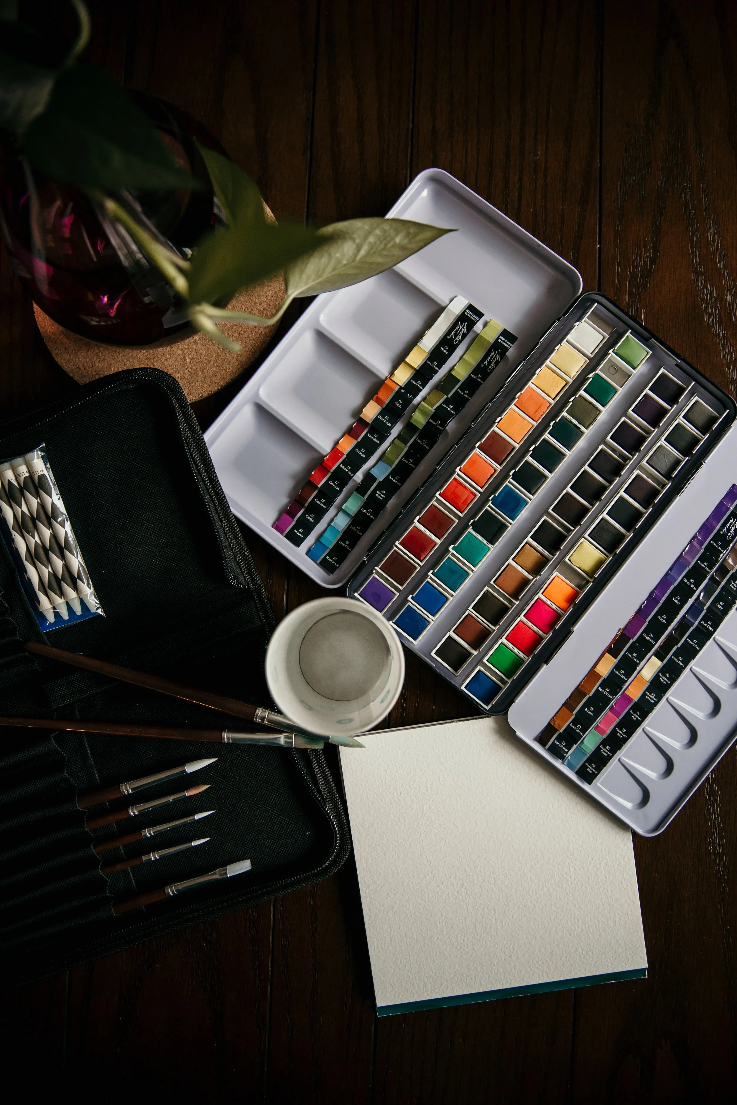 Artist's watercolor paint set, sketchbook, and art supplies on a dark wooden table. There is also a potted plant, a black pencil case, and a white mug.