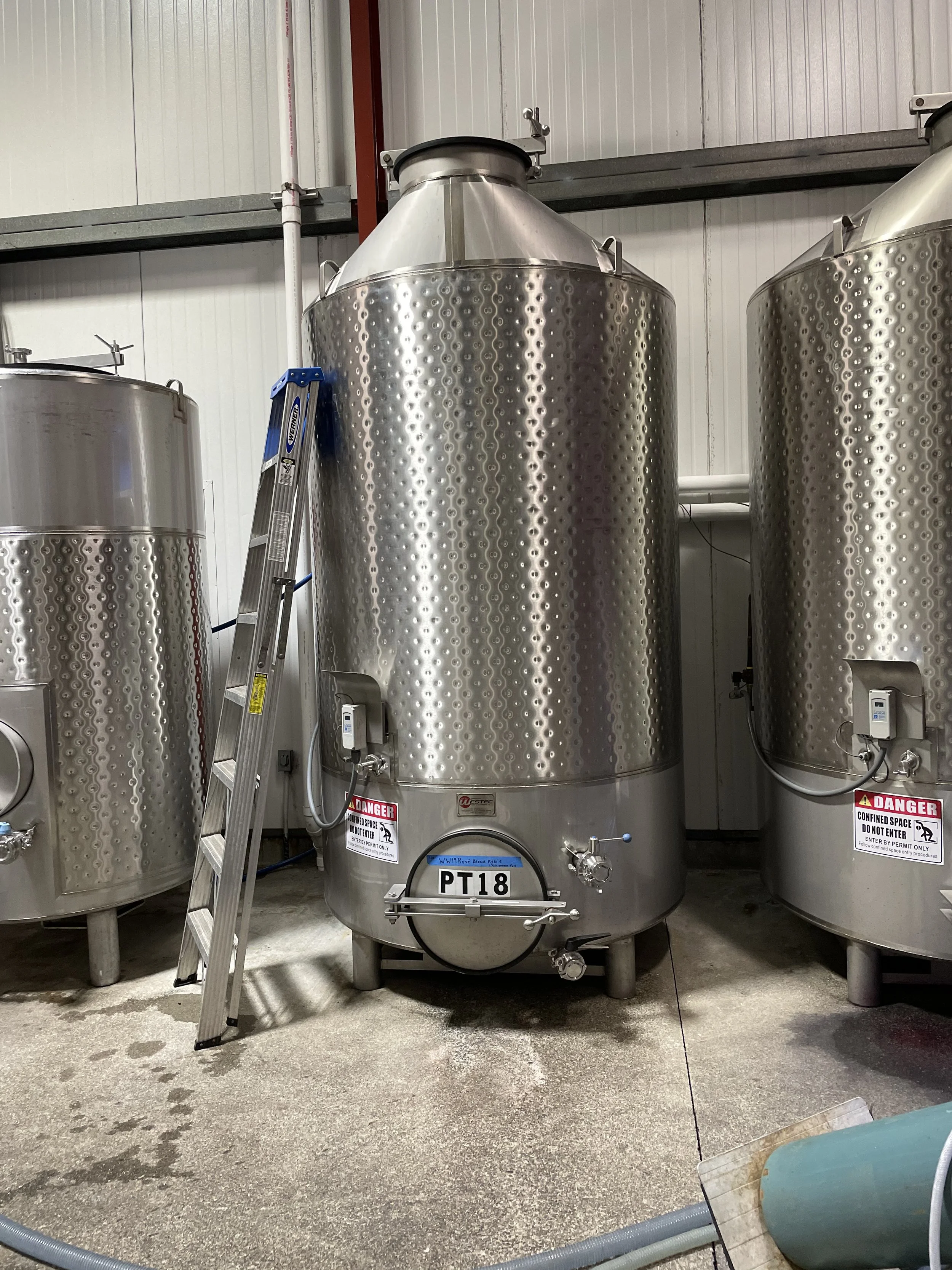 Large stainless steel fermentation tank in an industrial setting with a small ladder leaning against it and warning signs on it.