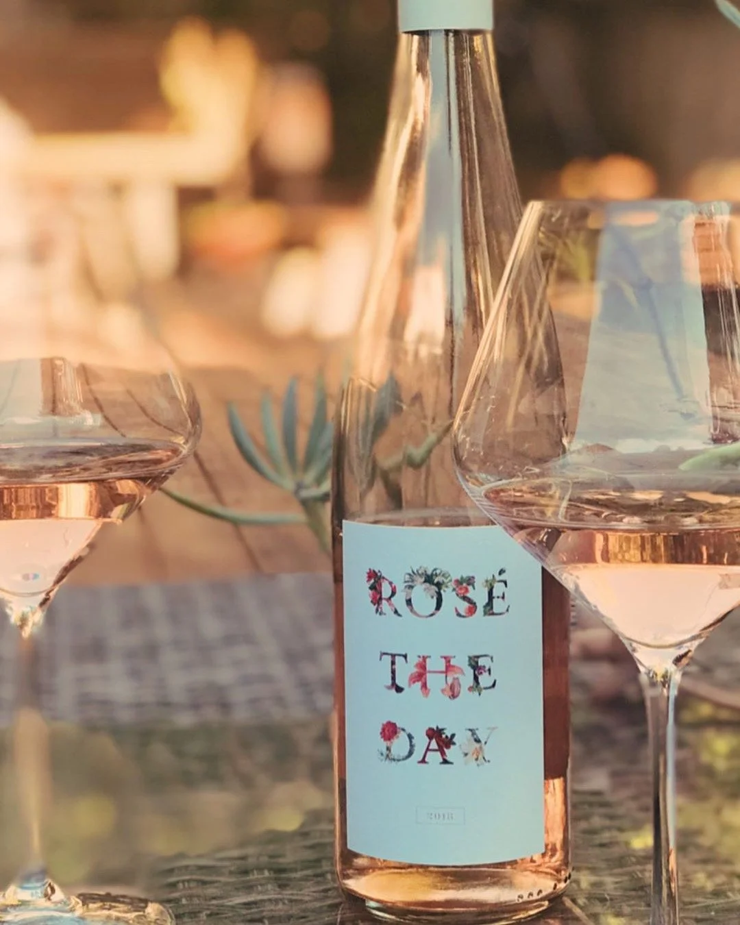 A bottle of rosé wine labeled 'Rose the Day' surrounded by two wine glasses filled with rosé wine, on a rustic outdoor table during soft sunset lighting.