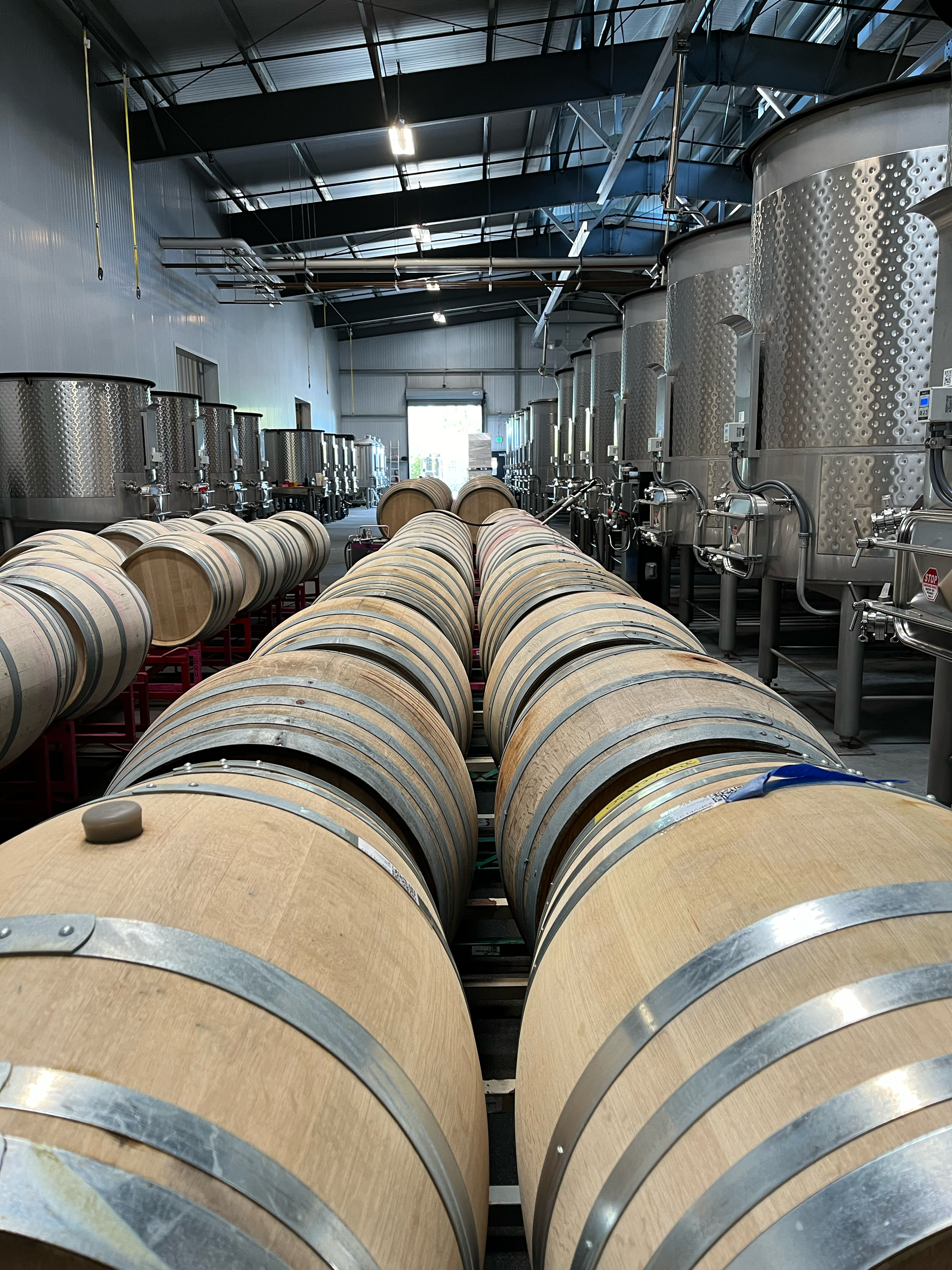 Storage tanks, possibly wine barrels, arranged in a row inside a warehouse or industrial facility with metal tanks and piping overhead.