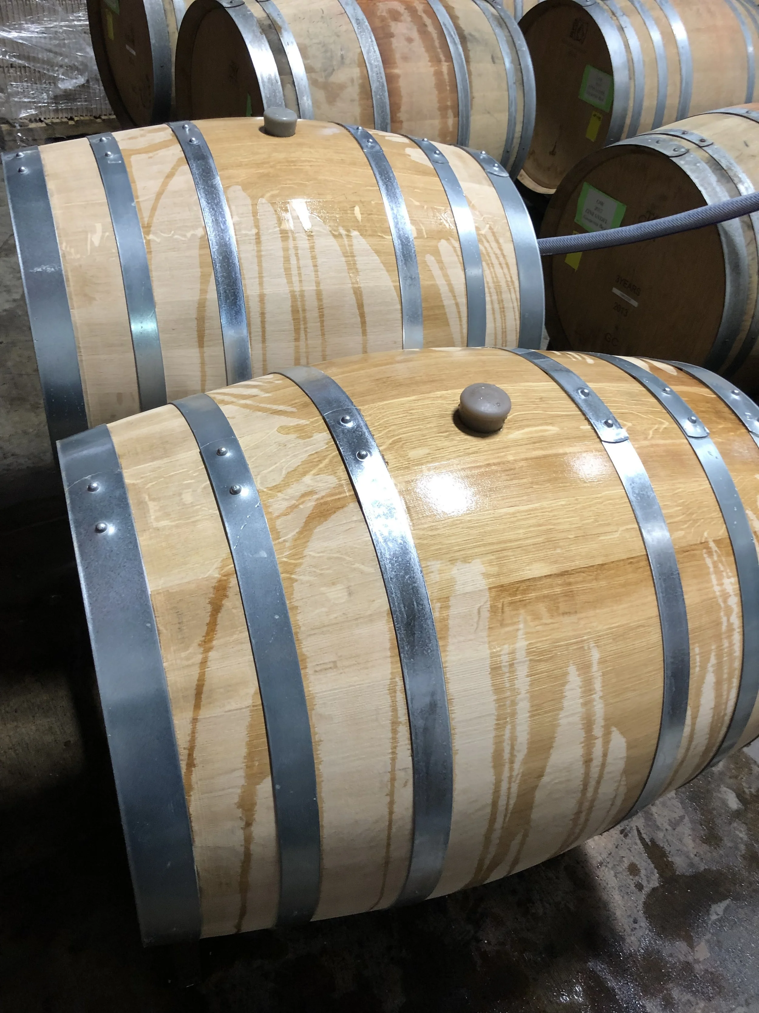 Large oak wine barrel with metal hoops and a wooden bung on top, placed on a concrete floor in a wine cellar or storage area.