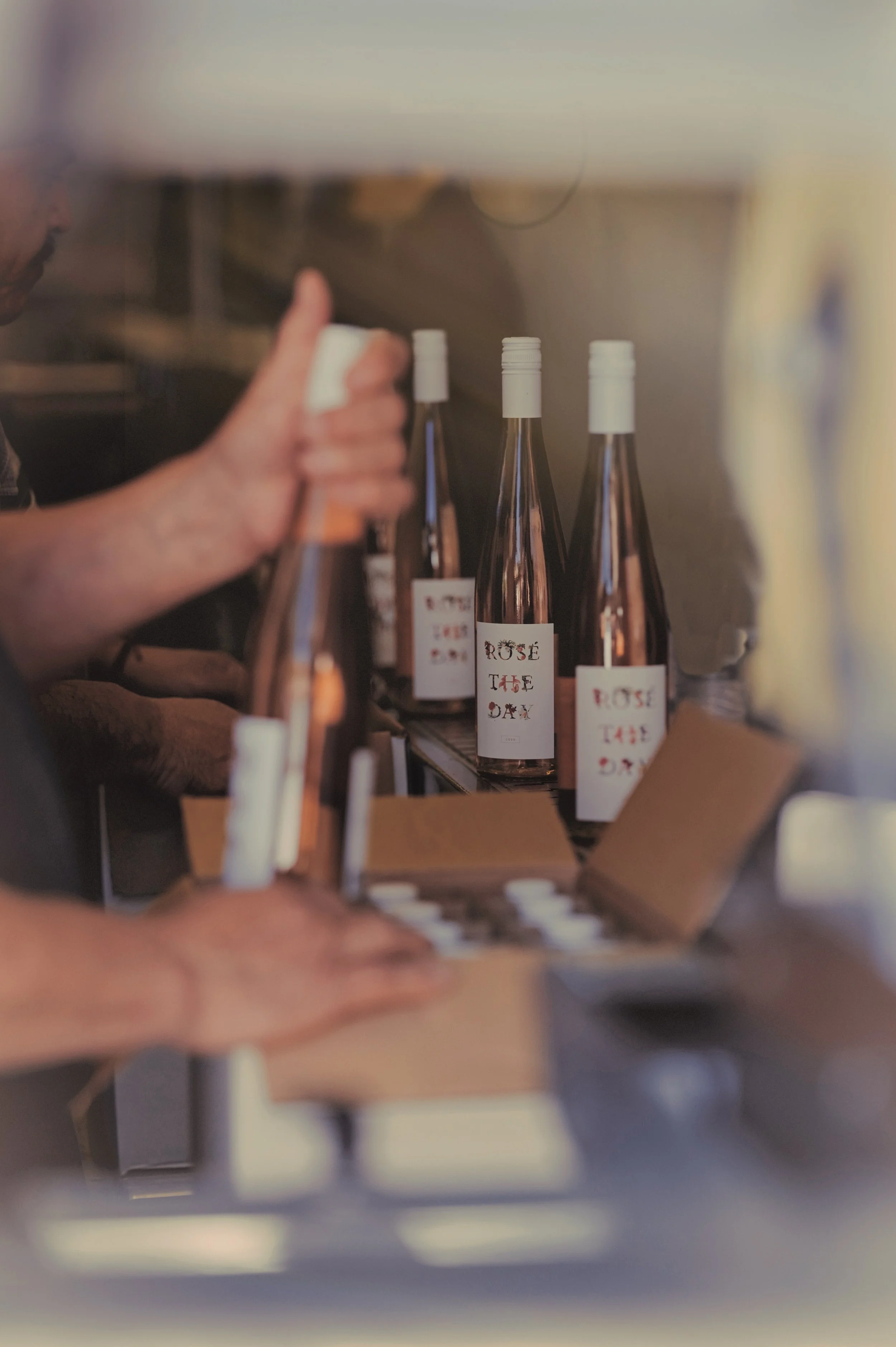 Close-up of a person reaching for a bottle of rosé wine on a shelf, with other rosé bottles with white caps and decorative labels visible in the background.