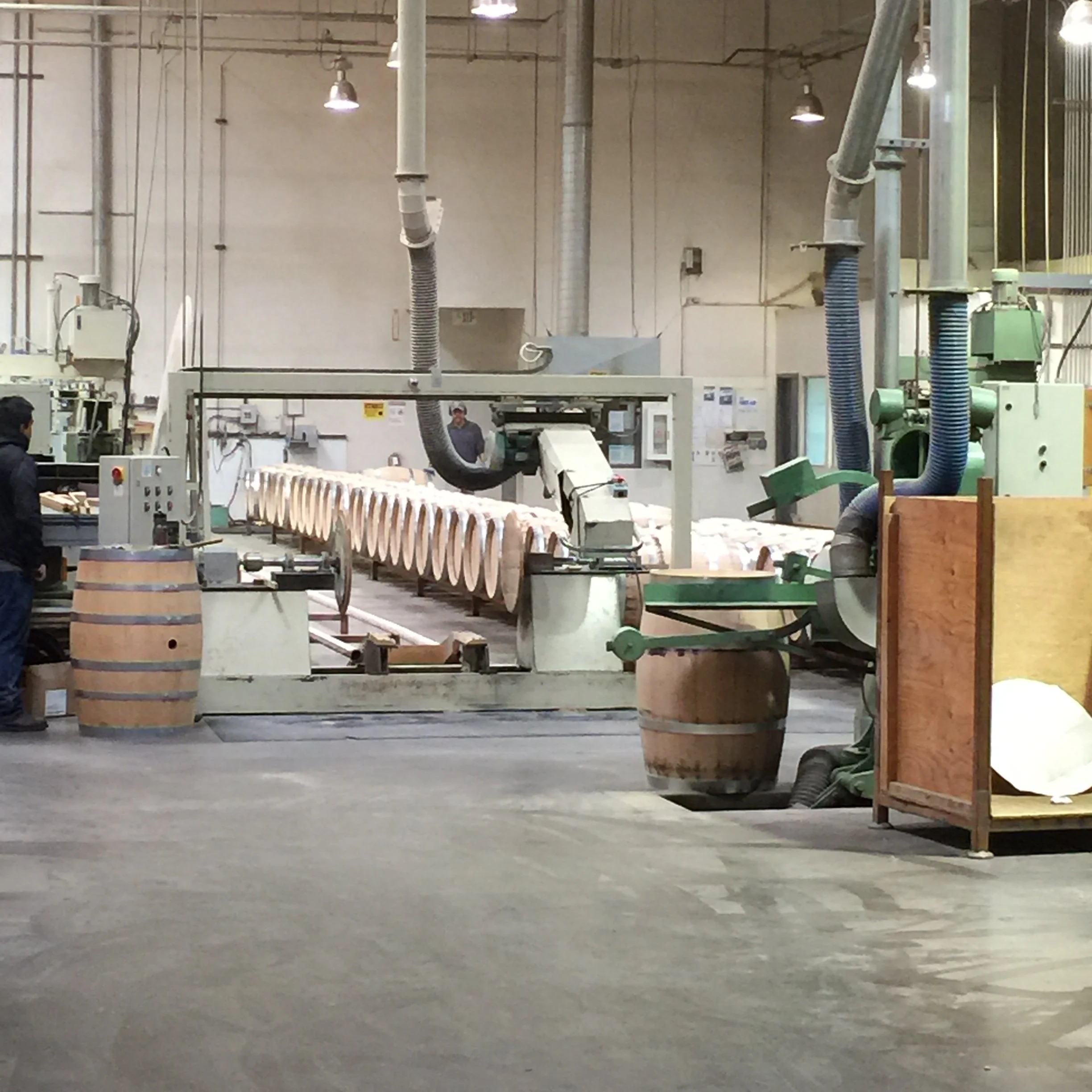 Large industrial workspace with machinery, ducting, and workers. The room has high ceilings with hanging lights and a visible wall in the background.