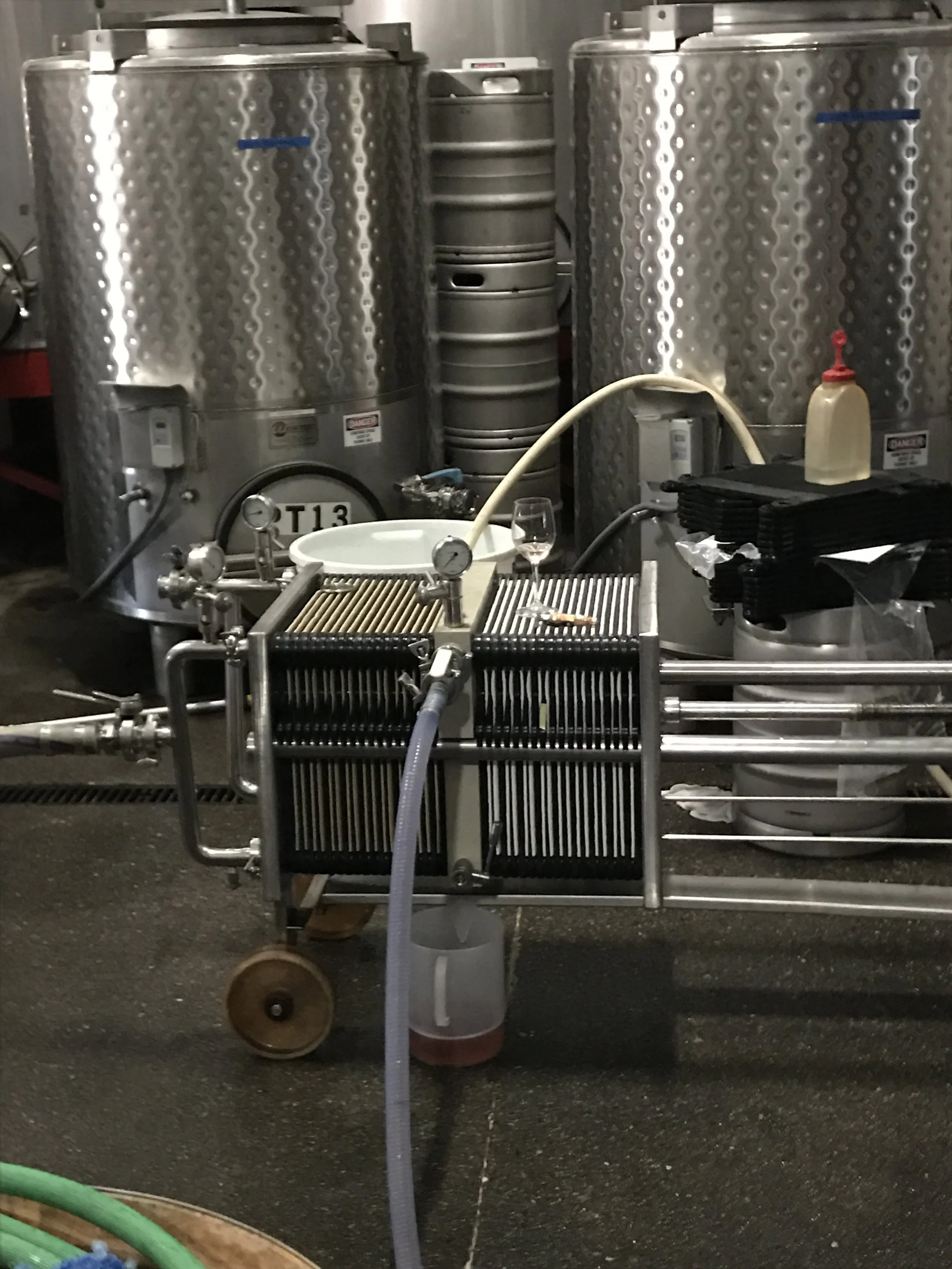 Two large stainless steel fermentation tanks with some stacked smaller barrels in the background, a tap, a clear glass with red wine, and a bottle of sanitizer, all in an industrial setting.