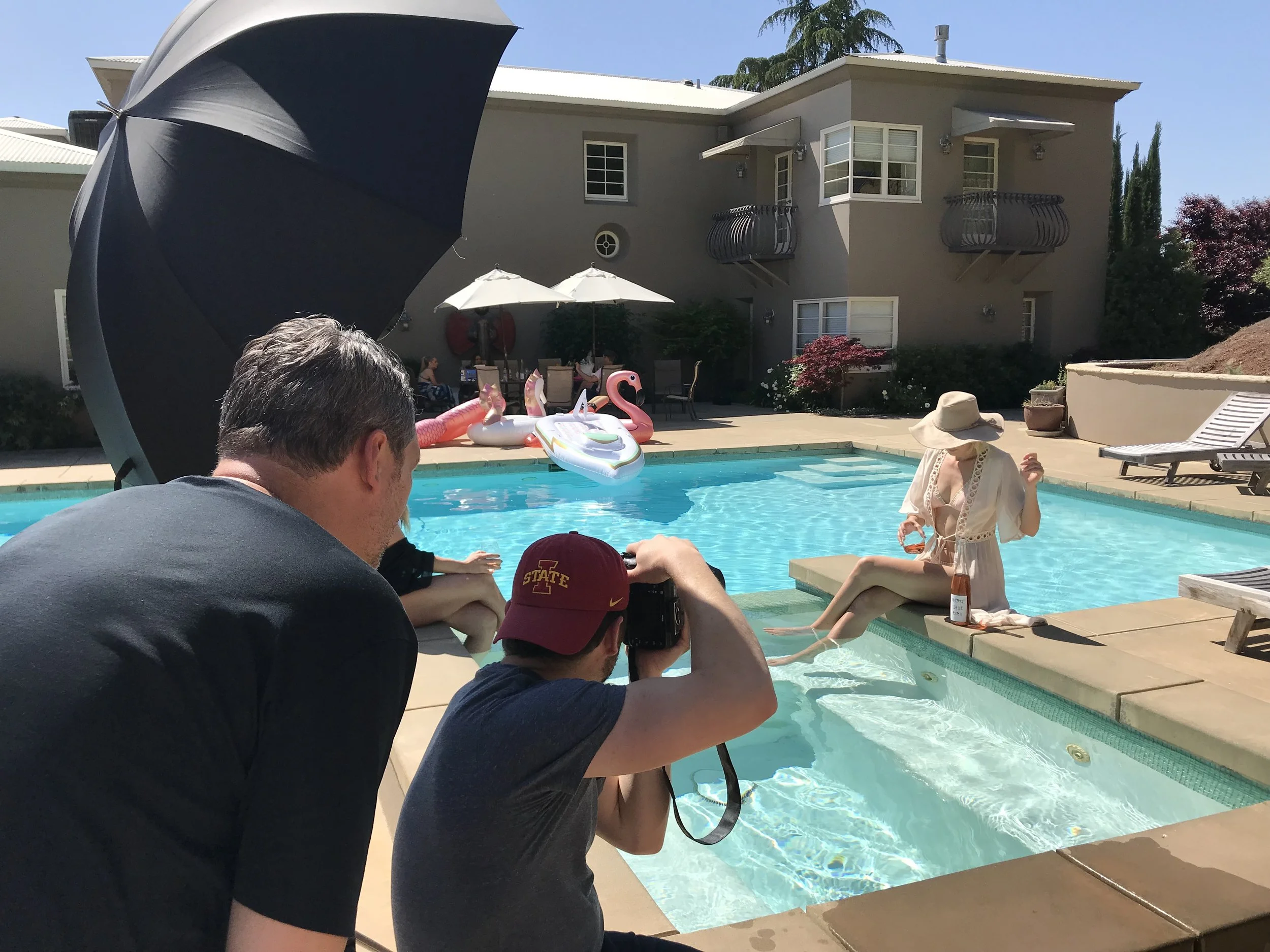 People sitting and standing by a backyard swimming pool with floating flamingo and unicorn pool toys, umbrellas, and a modern house in the background.