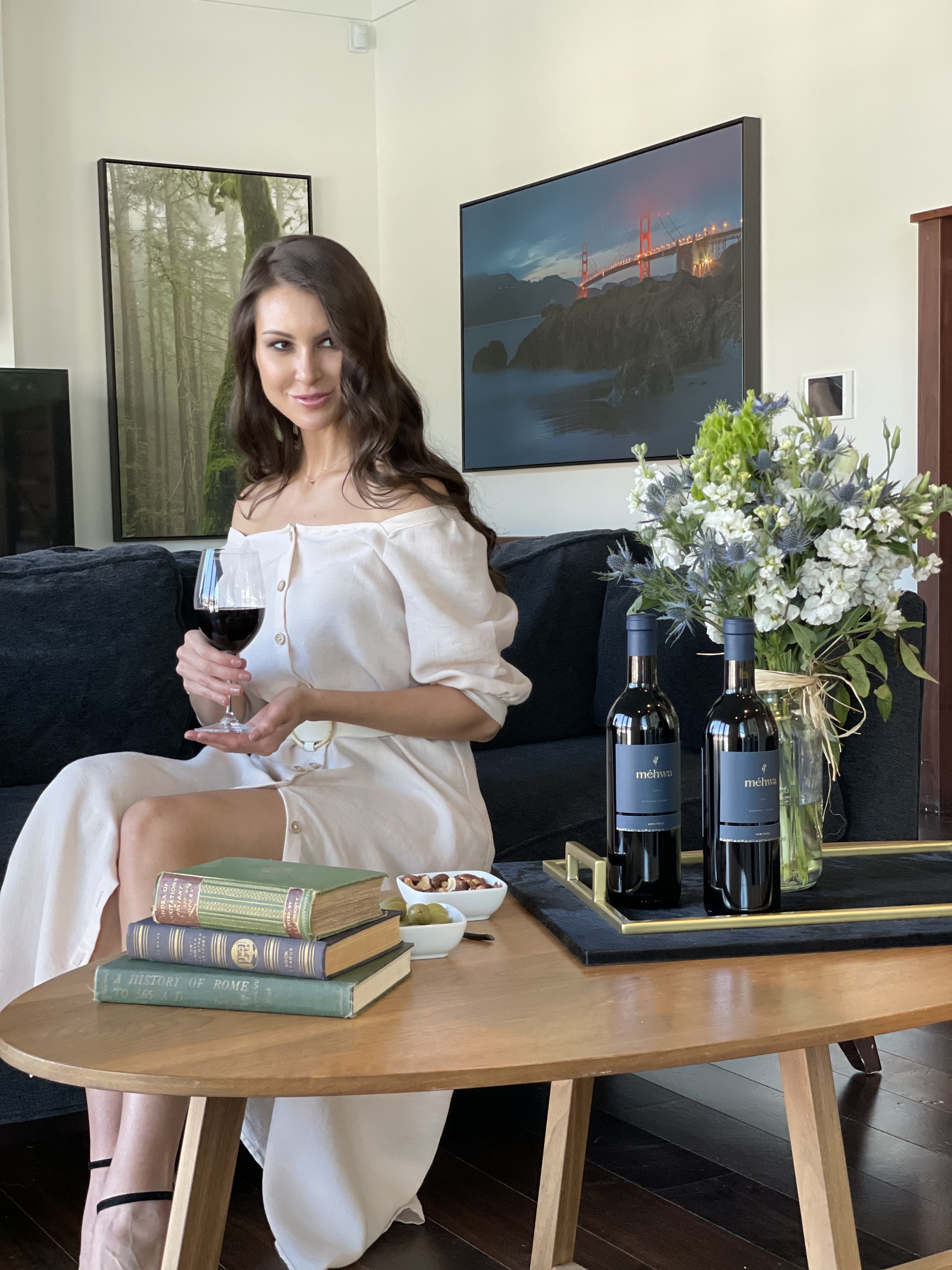A woman with long brown hair and light skin, wearing a beige dress, sitting at a wooden table in a living room, holding a glass of red wine, with a flock of books, a flower arrangement, and some food on the table.
