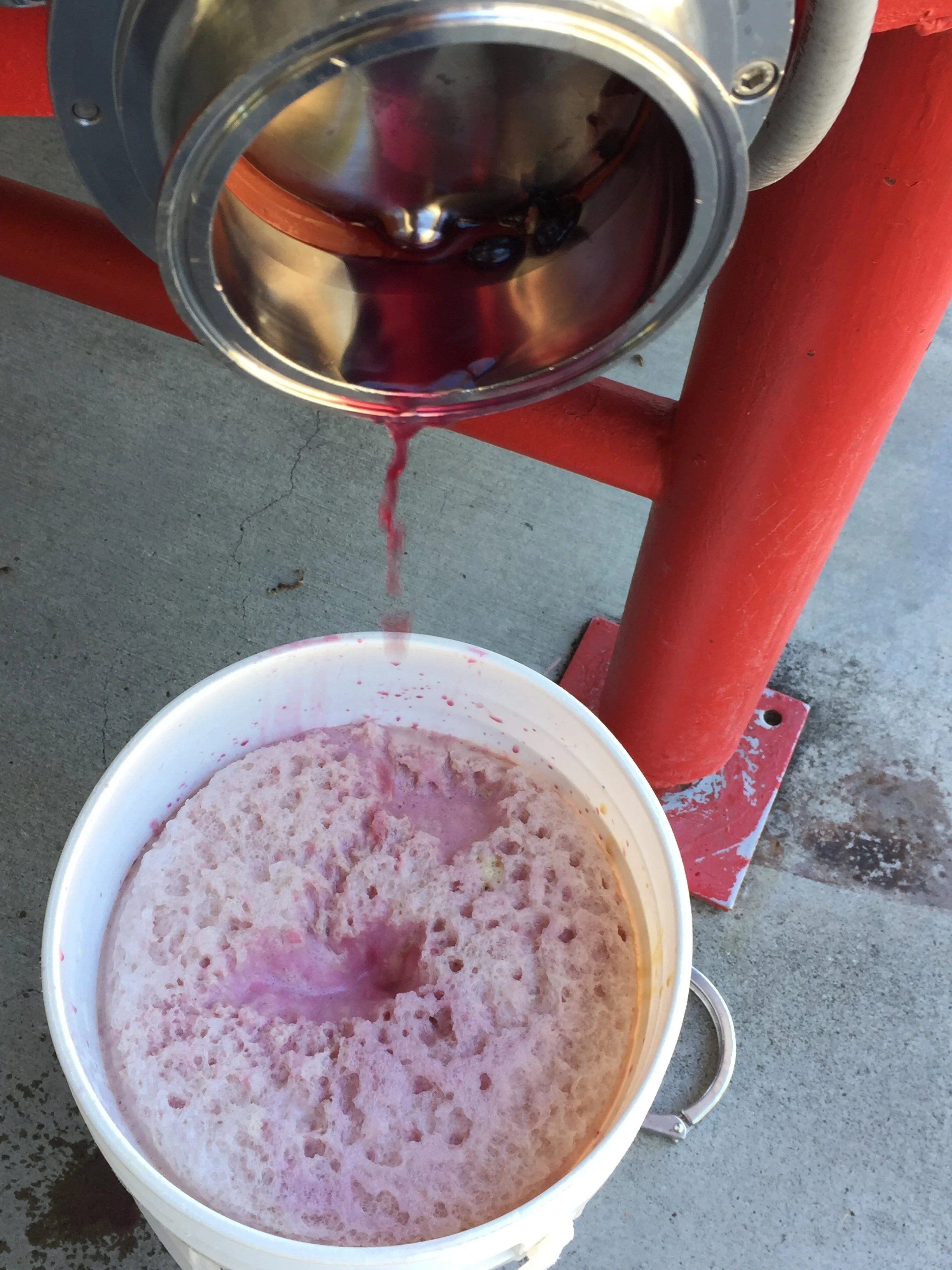 A bucket with a frothy, pinkish liquid and a red stain dripping from the spout, positioned on a concrete surface with a red metal structure and gray pipe in the background.